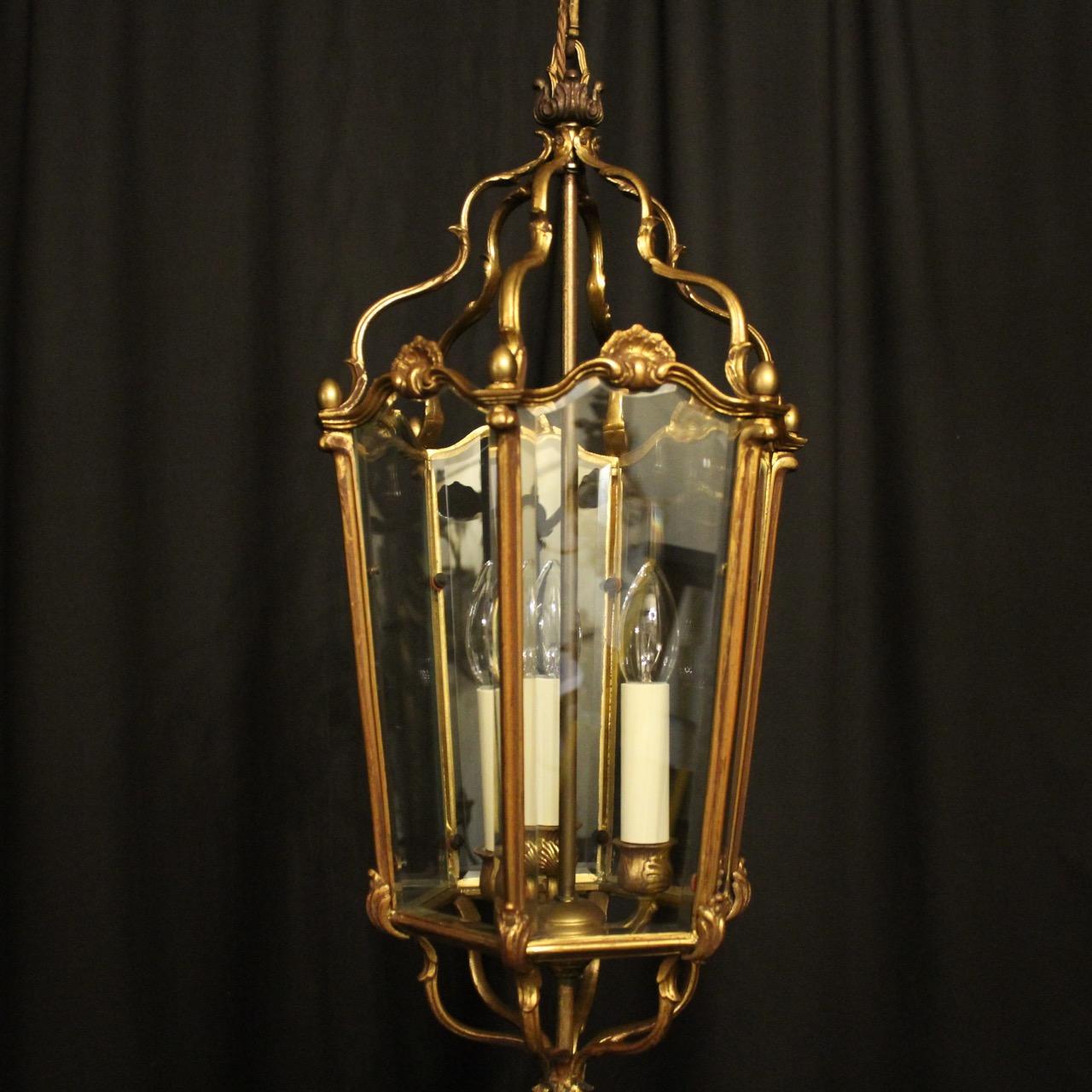 A French gilded bronze triple light hexagonal antique hall lantern, the six bevelled tapering glass panels held within an ornate scrolling framework with three light fittings and having decorative scrolled central shell embellishments, with edged