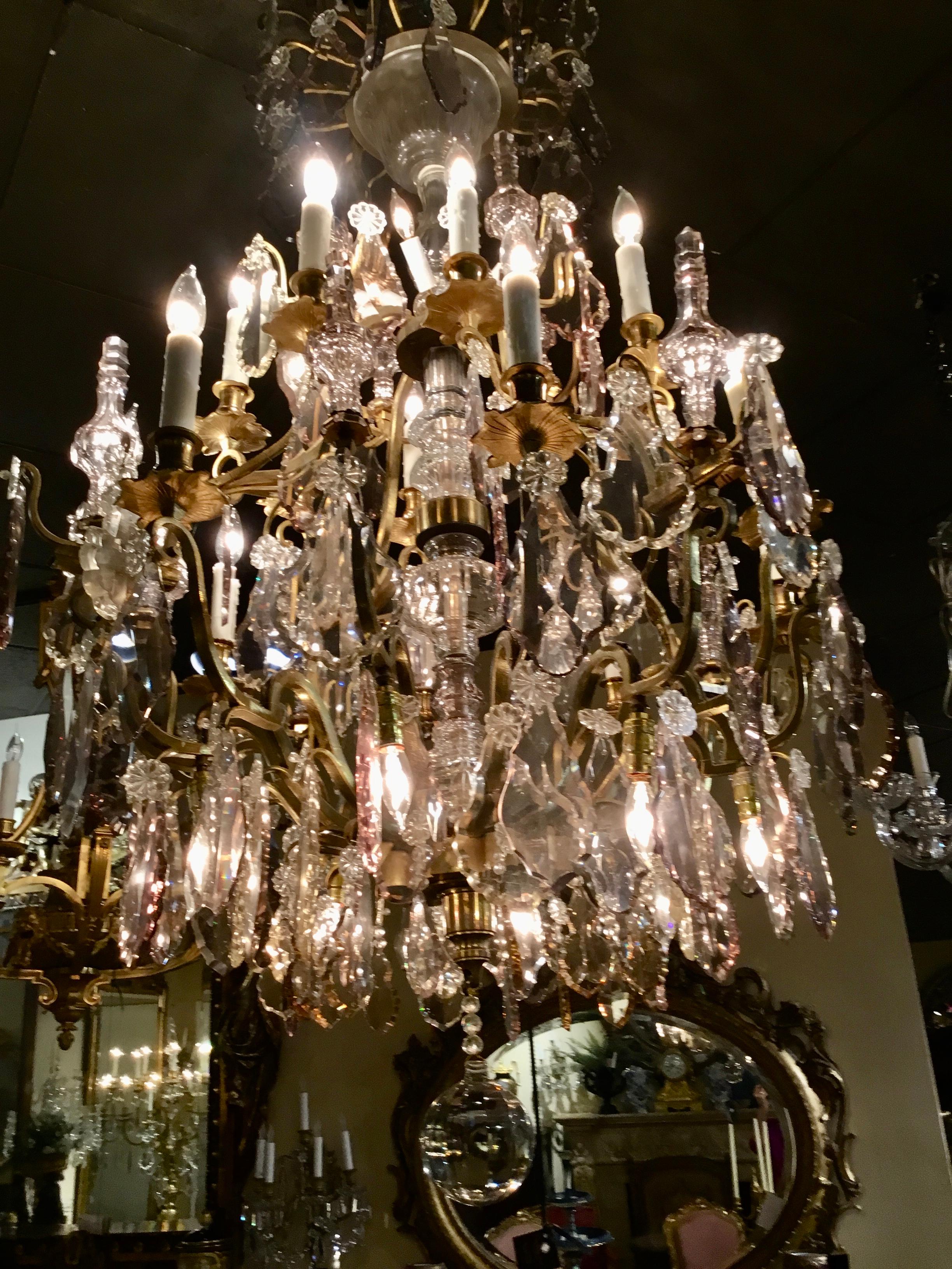 Gran y brillante araña francesa dorada de bronce de 28 luces. Bronce dorado y poste central con 
Brazos en forma de volutas con un gran cristal transparente y cristales dispersos de color lavanda muy pálido.
Hay remates de cristal en todo el