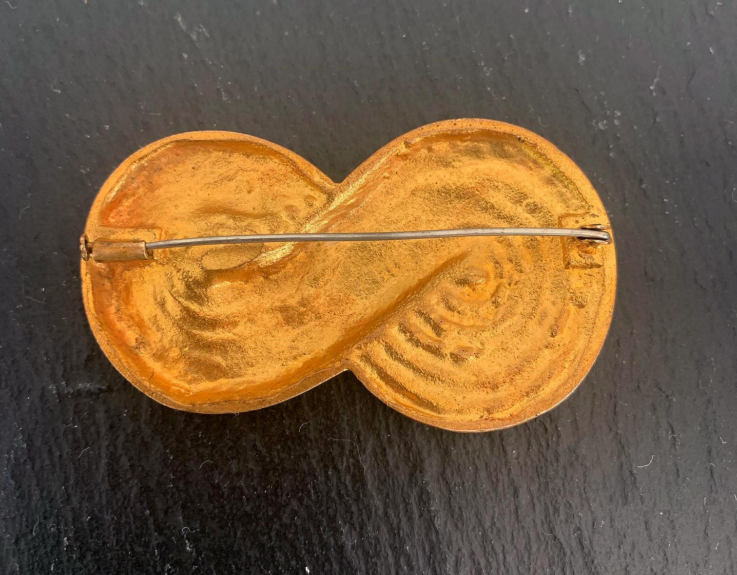 A brooch in gilded bronze by French art jeweler Line Vautrin circa 1950s-1960s. The unusual model depicts the mathematical sign of infinity, on which appears in relief inscription of the extract of L'Enfer by Dante Alighieri (1265-1321). 