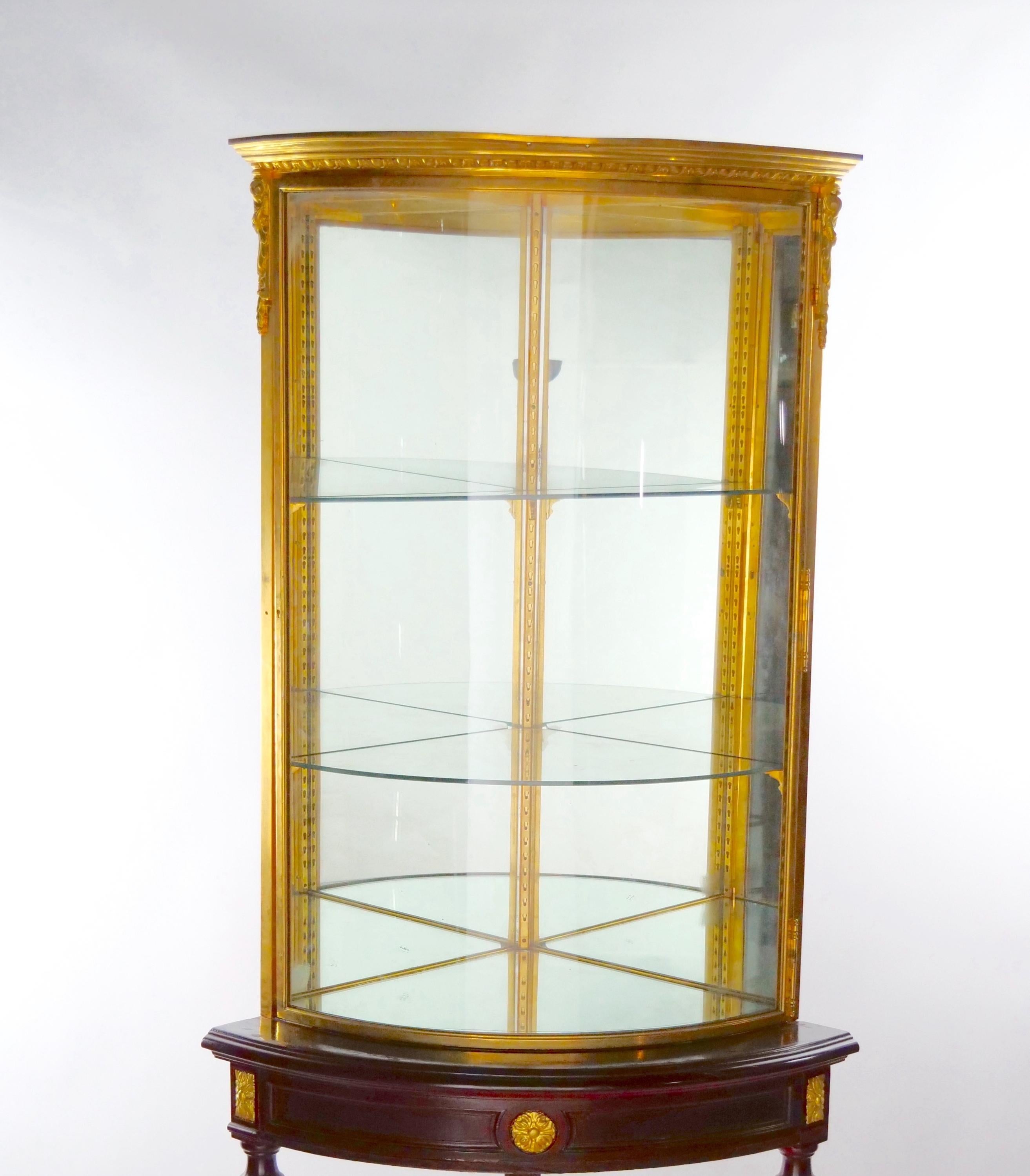 French two piece gilt bronze mounted and glass frame with gilt accent mahogany wood frame holding base curio cornered display cabinet. The curio / vitrine cornered cabinet features a demilune shape, gilt wood legs with a Y shape stretcher bar with a