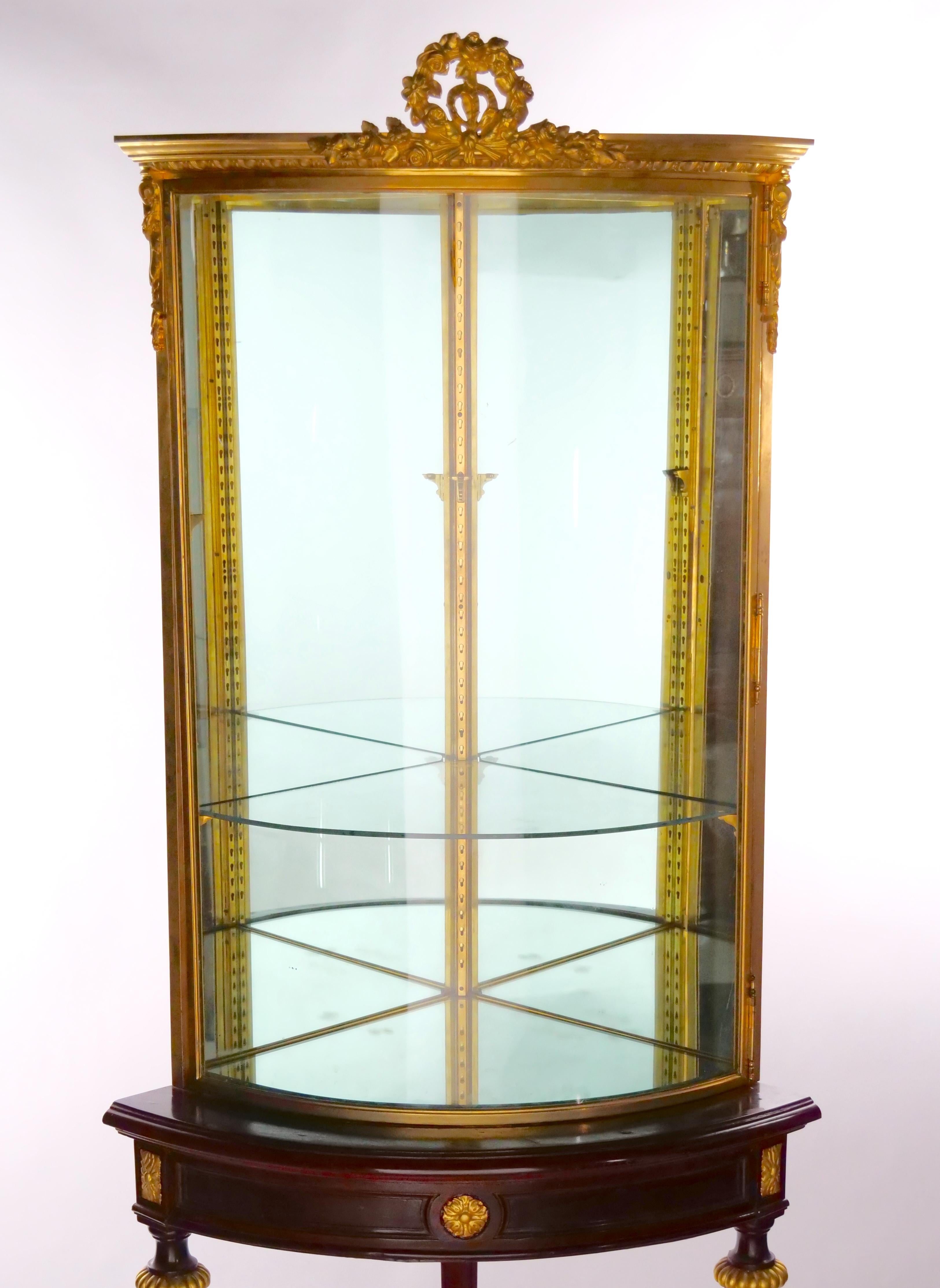 French two piece gilt bronze mounted and glass frame with gilt accent mahogany wood frame holding base curio cornered display cabinet. The curio / vitrine cornered cabinet features a demilune shape , giltwood legs with a Y shape stretcher bar with a
