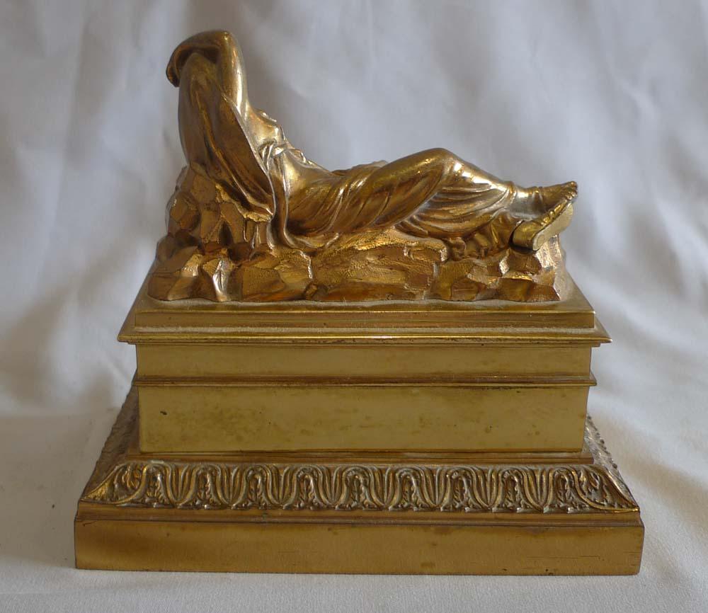 French inkwell in gilt bronze. Classical lady after the antique to the lid. She lying back against a rock with sandals and long robes and her arm over her head. To the inkwell front is an applied gilt bronze mount of palm fronds. The base has a fine
