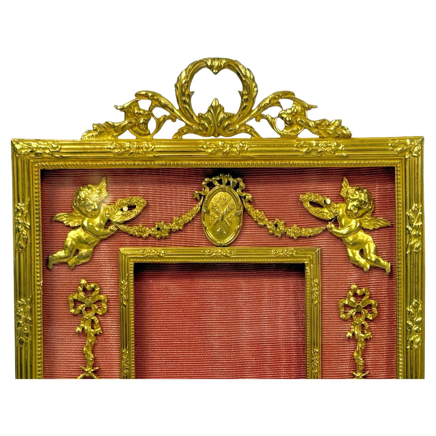 Cornice fotografica classica francese in bronzo dorato. La cornice presenta decorazioni in Ormolu con Putti, nastri, fogliame e ghirlanda con medaglioni. I pezzi dorati sono adagiati su un tessuto di seta rosa Fabrice. L'apertura centrale della