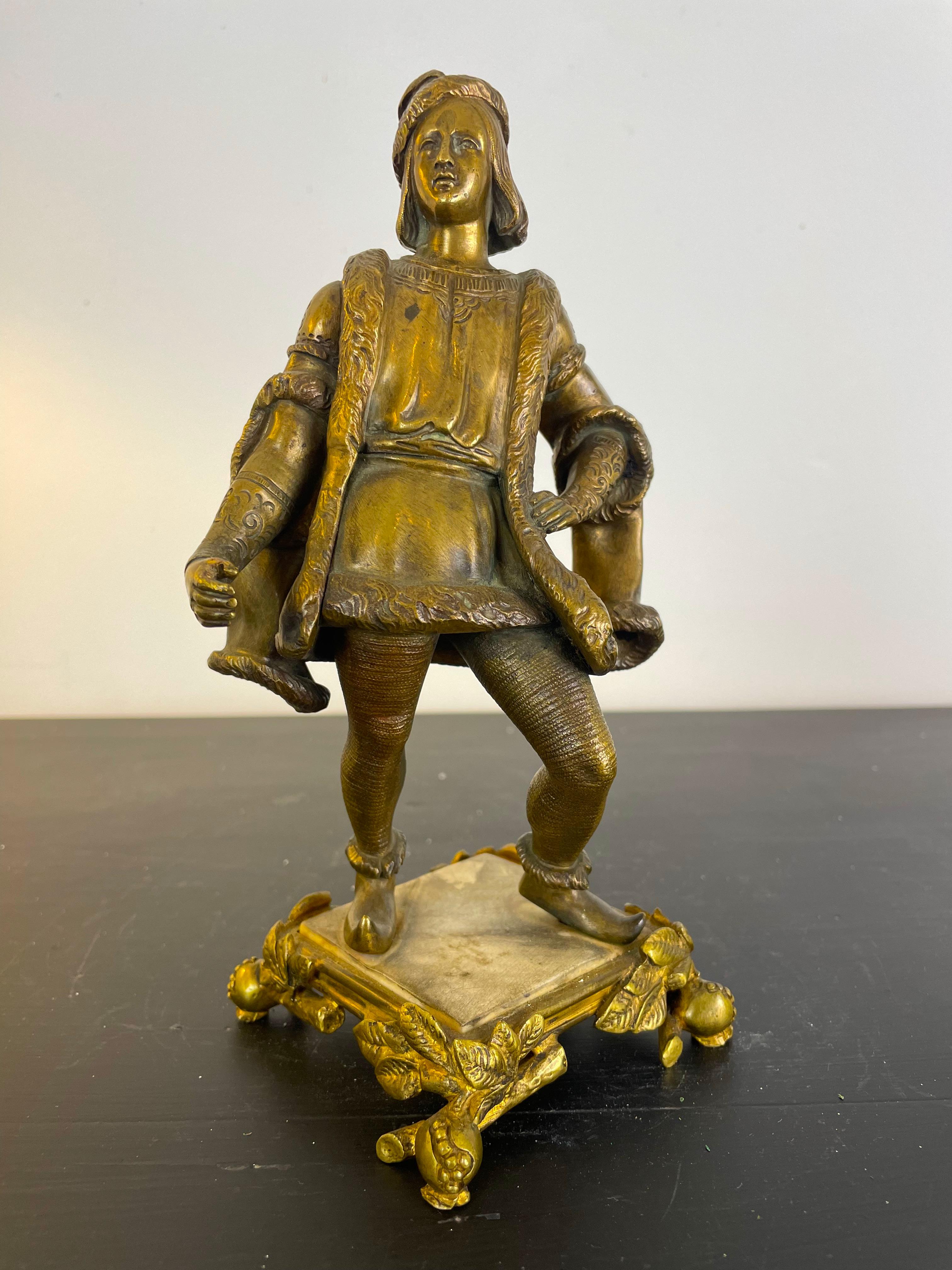 Sculpture en bronze doré représentant un jeune homme galant en costume Renaissance, dans le style du roi français Henri IV. Il est vêtu d'un doublet pour le haut, et par-dessus il porte un manteau de style casaque, porté largement ouvert, dont les