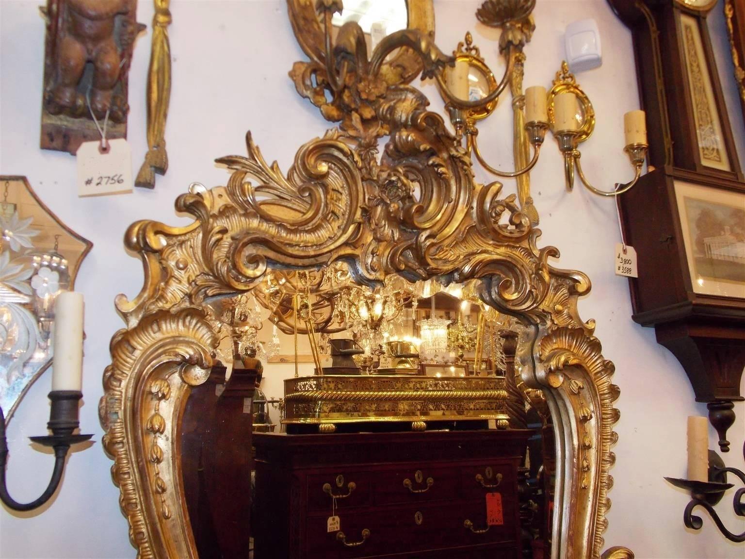 French gilt carved wood foliage wall mirror with a centered cartouche, sweeping scrolled sides with graduated bell flowers, flanking flower baskets, and terminating on a decorative floral border. Mirror retains the original beveled glass and wood
