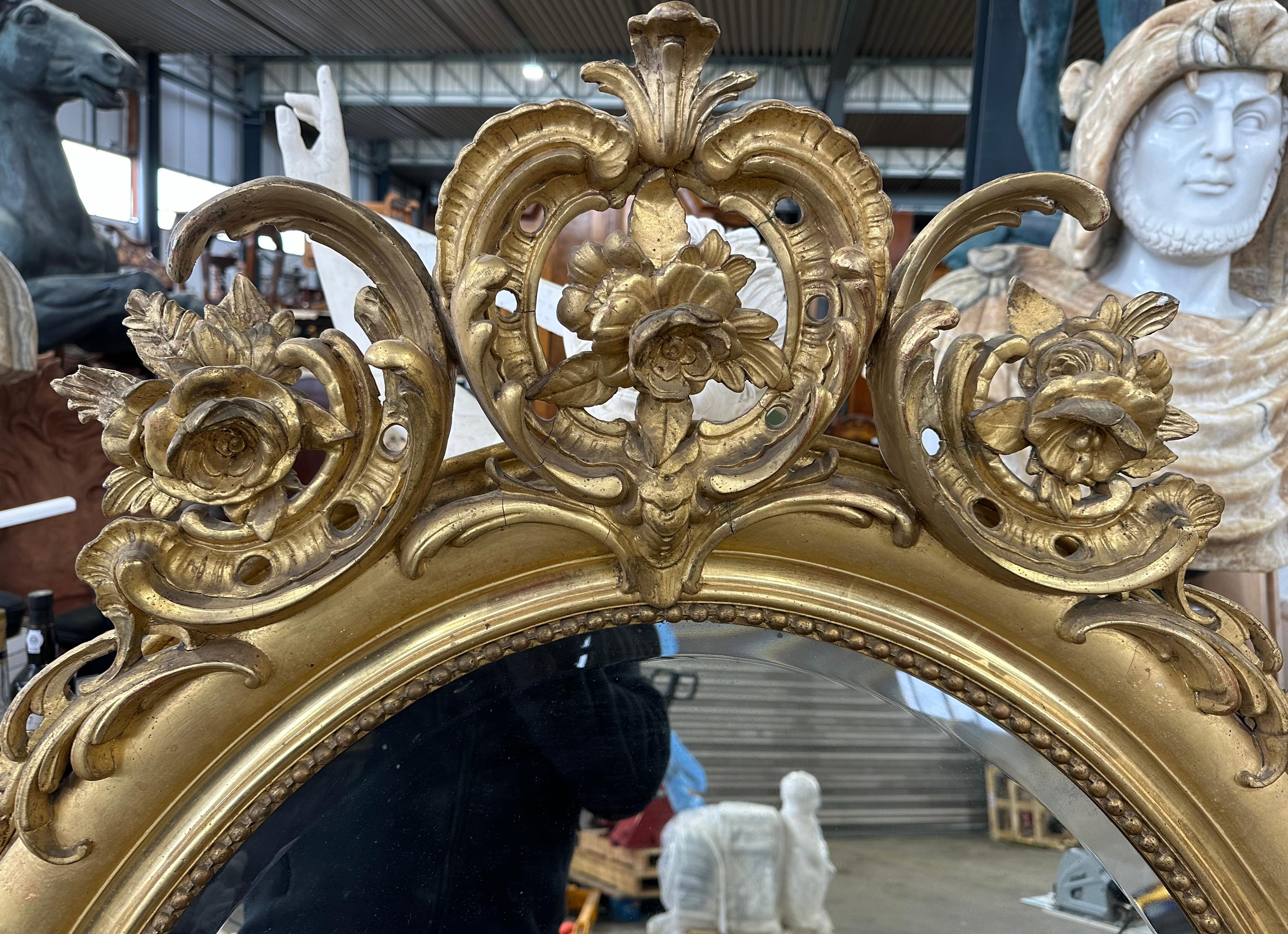 Miroir décoratif français en bois doré avec des sculptures florales et une crête élaborée avec trois panaches au sommet formés de rinceaux et de feuillages. Le verre ovale est biseauté avec des perles dorées. Un miroir très attrayant qui donne une