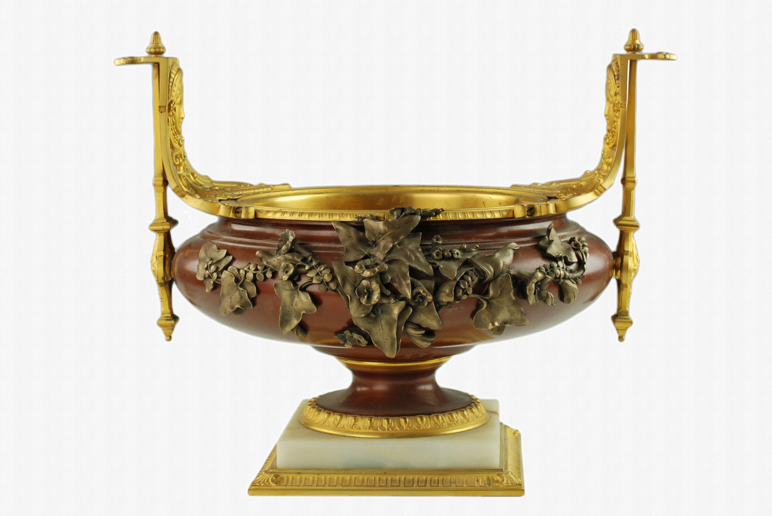 This exceptional late 19th century three-piece French garniture includes two footed urns and a low footed centerpiece bowl. The urns and bowl are composed of patinated bronze with ormolu mounts which include neoclassical mask detail and rest on