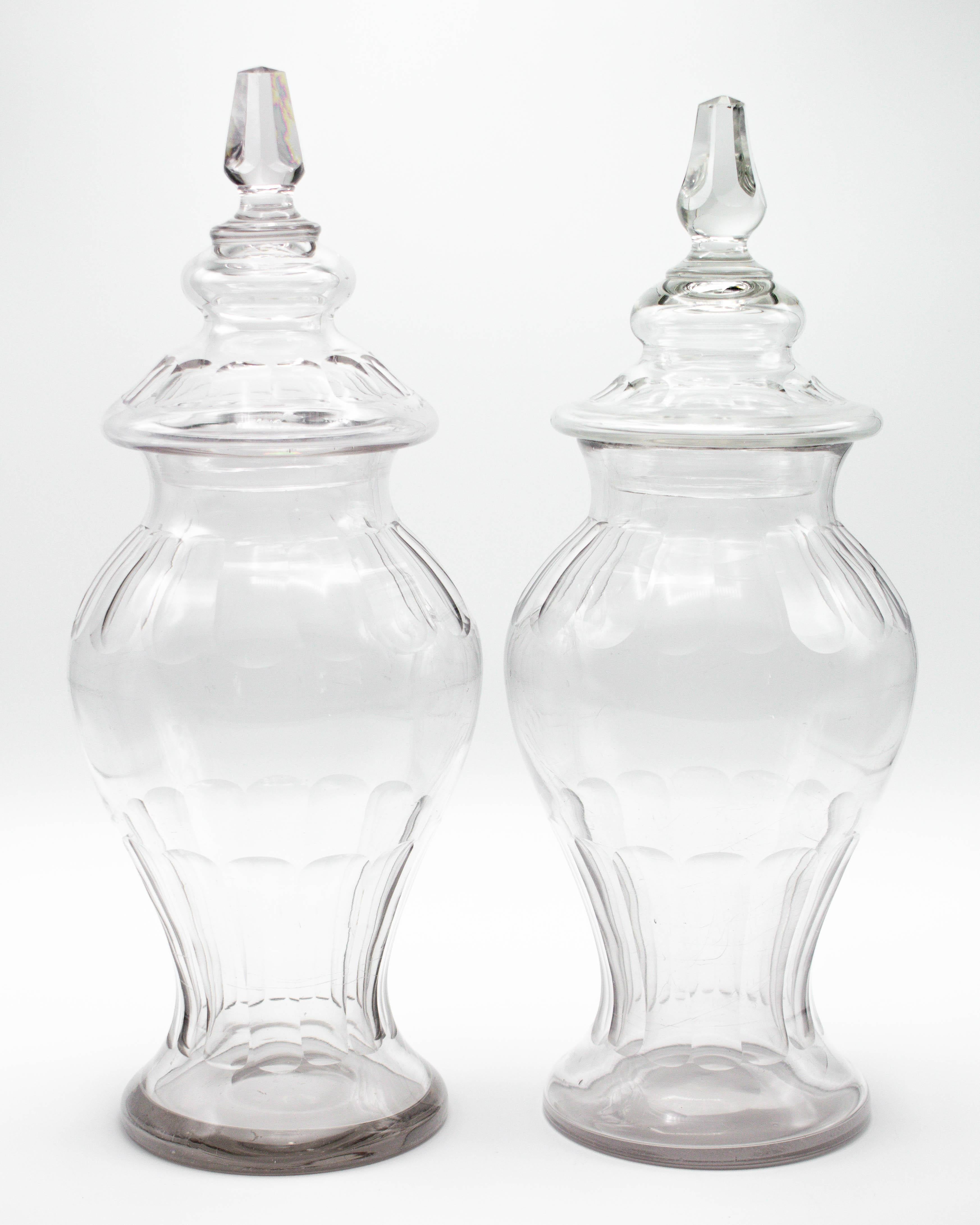 A pair of French glass lidded apothecary or candy jars. Urn shaped faceted form. Lids with cut glass knobs. In good condition for the age and use, with light scratches and small chips to knobs. Purchased from a confiserie, or sweet shop, along many