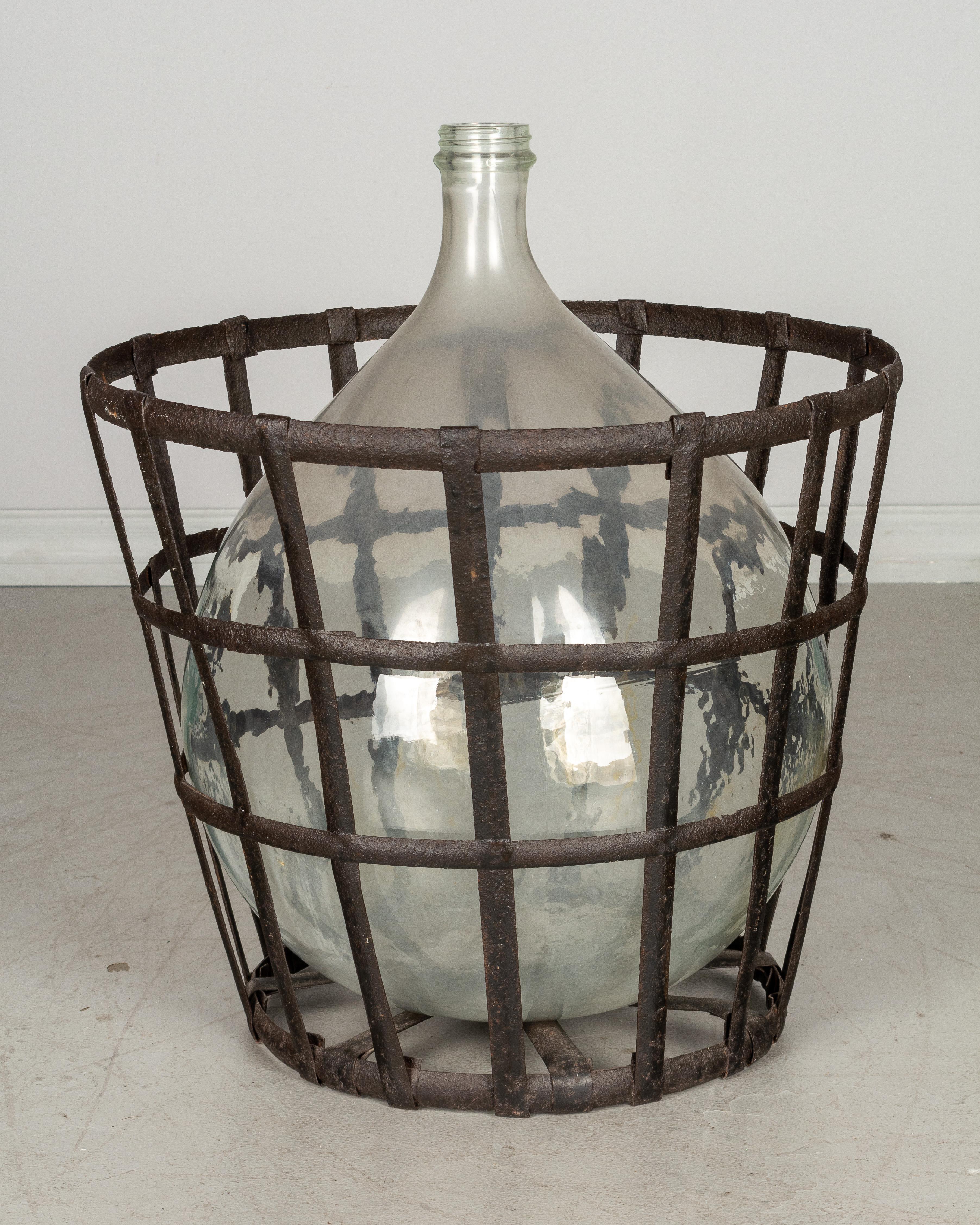 A large French globular form glass dame Jeanne, or demijohn, bottle in original metal cage basket with weathered patina. Capacity: 25 liters. Weight: 16 lbs.