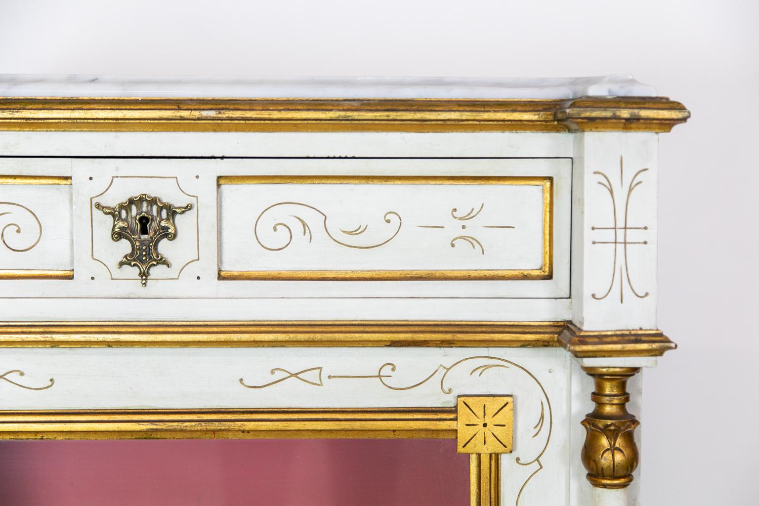 This display case has an ogee shaped Carrera marble top framed by gold molding. The frieze has two recessed panels with incised gold stylized arabesques. The front stiles have two support fluted columns that have carved tops and bases. The front