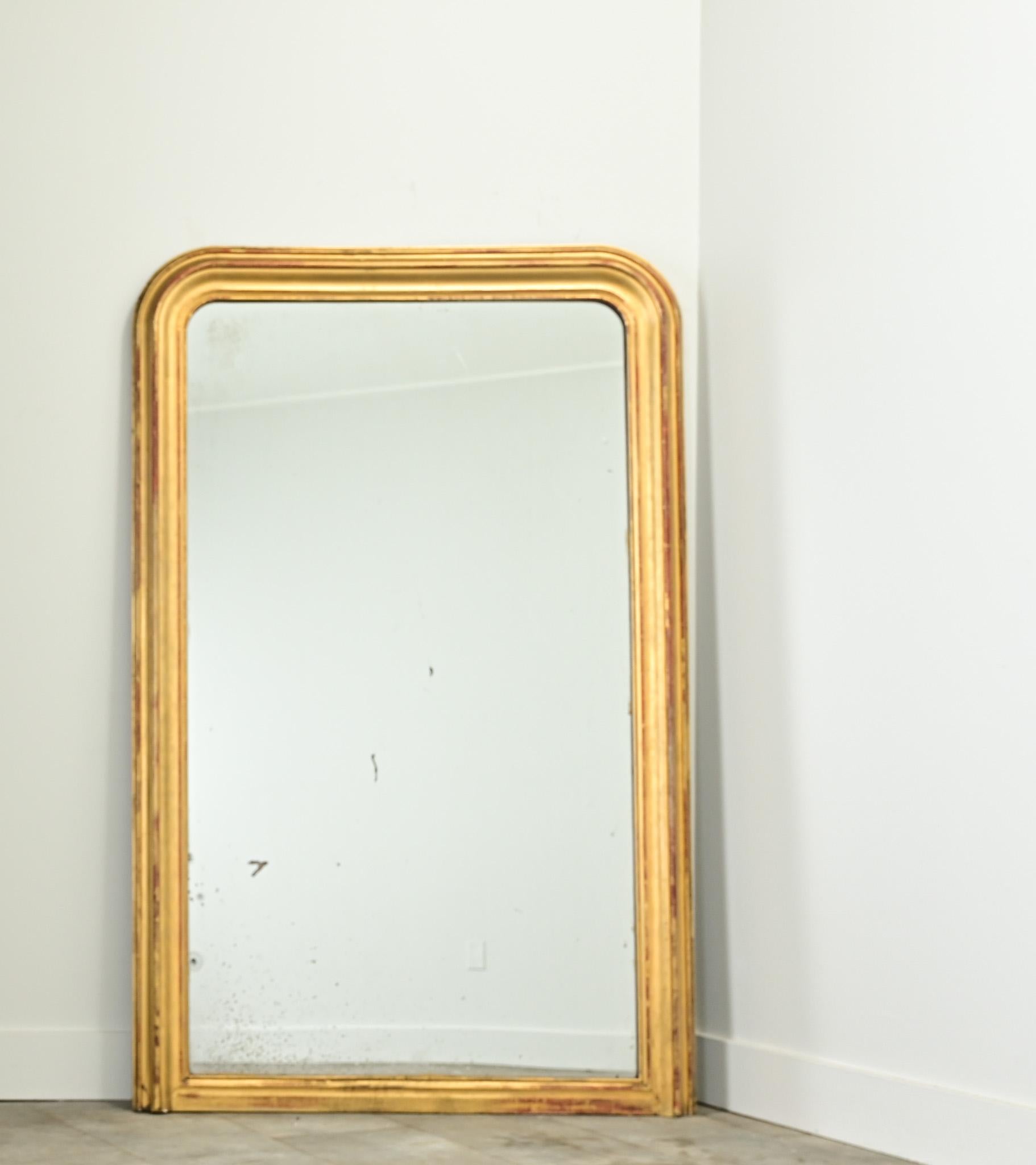 A French gold gilt Louis Philippe mirror from the 1800’s. The simple molded frame has rounded corners at the top and its original gold gilt finish, revealing the reddish bole beneath. The mirror plate is original and has light foxing and