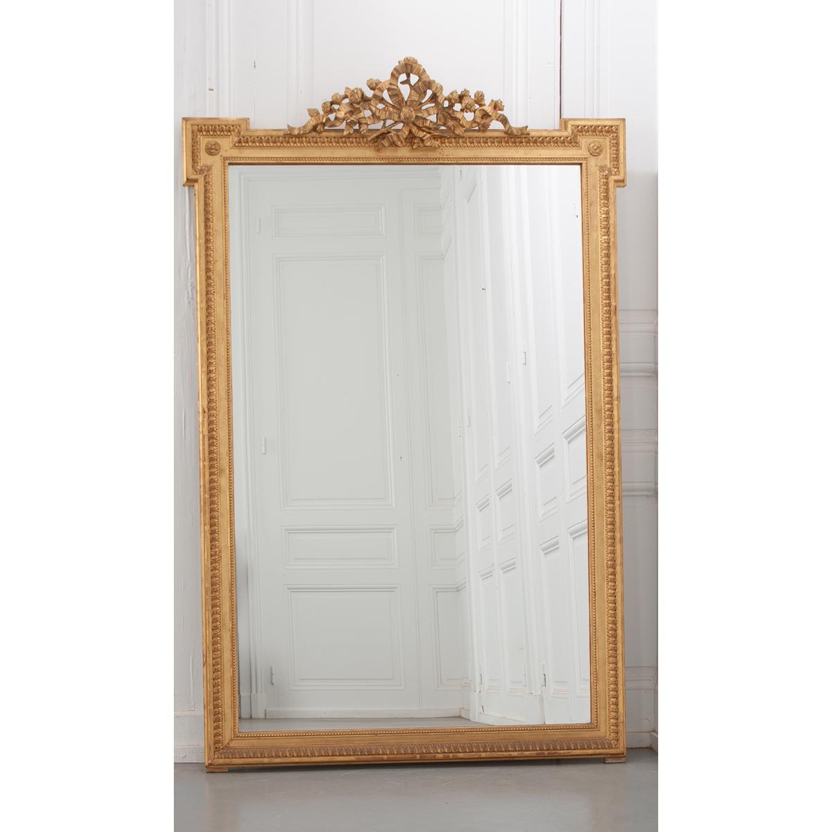 A French gold gilt Louis XVI style mirror with its original mirror plate. It has some aging. The crest is achieved with a large bow in the center and flowers entwined in the bow. It has a decorative edge. Be sure to view the detailed images.