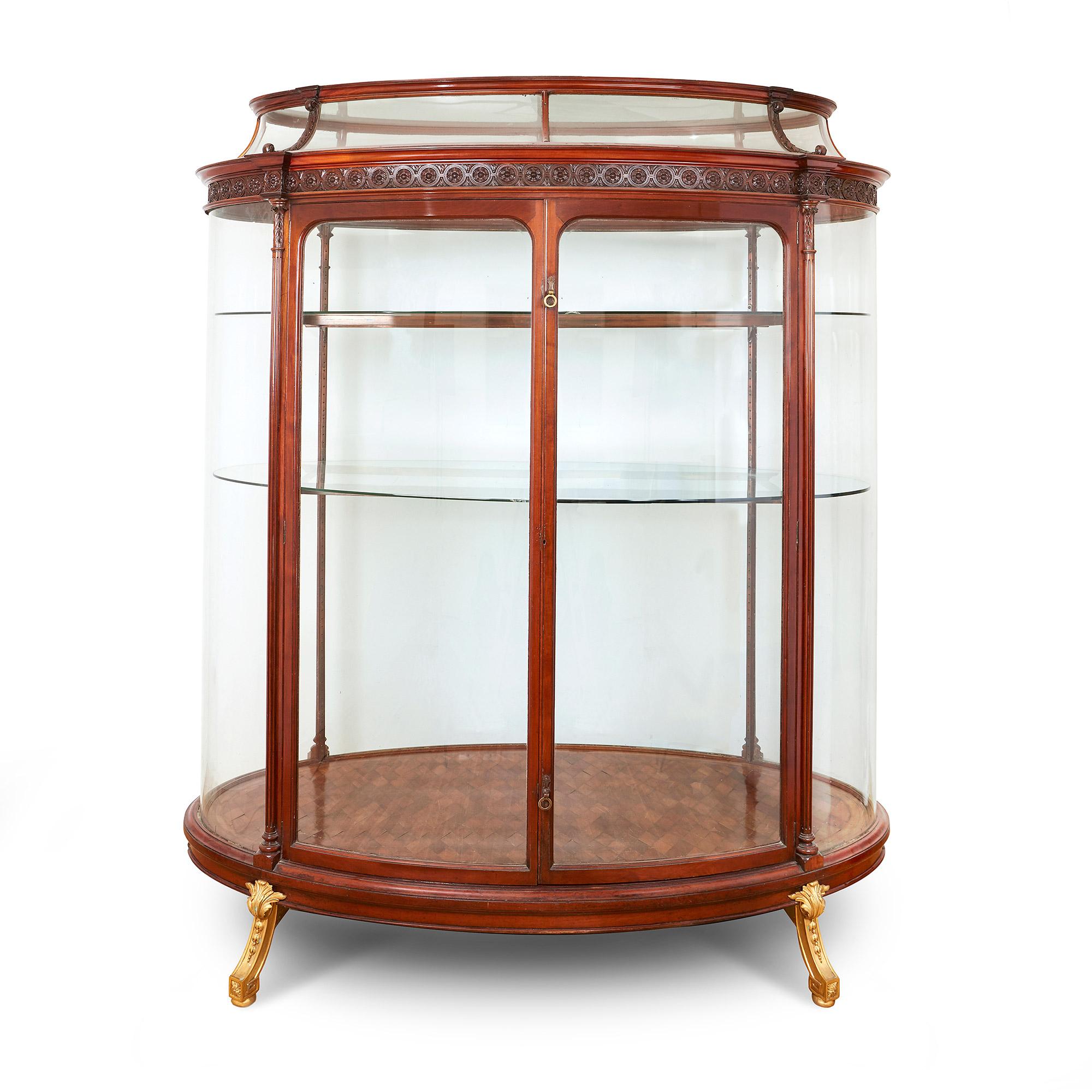 The vitrine oval shaped, with central hinged tall doors to the front and glazed display all around, mounted with ormolu handles and short legs.