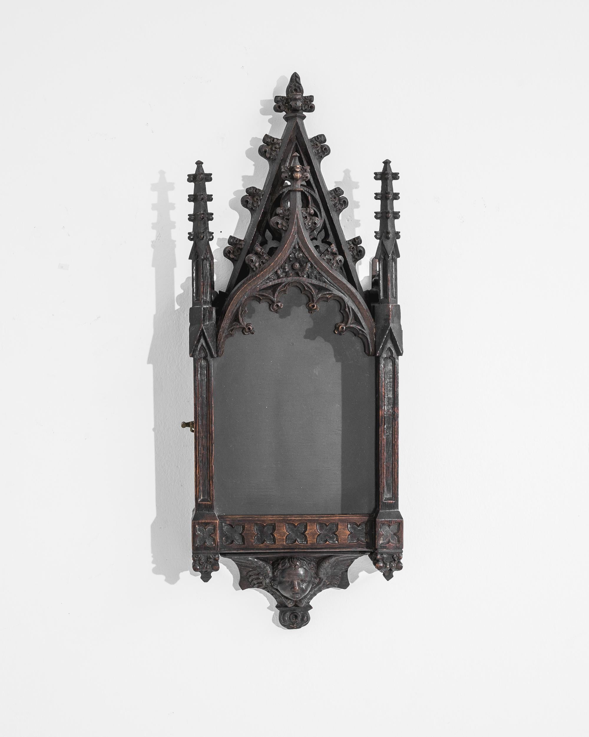 A small wooden display cabinet from 1900s France, this antique find flaunts an outstanding gothic inspired profile. Two solemn pillars flank an acute pinnacle, crowning a central glass door garnished with hand carved ornamentations. A frieze of