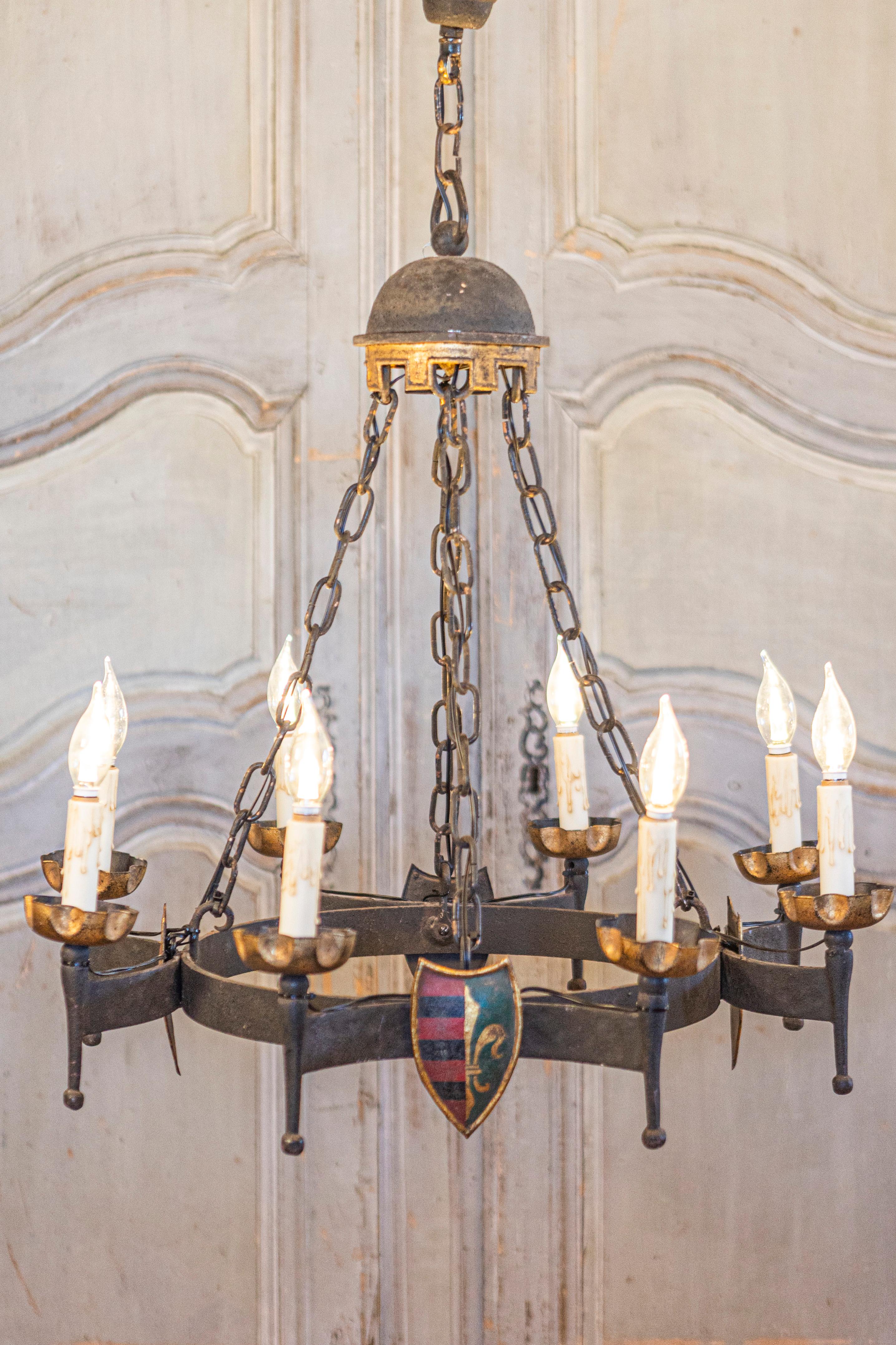A French Gothic style eight-light iron chandelier from the 20th century with hand-painted crests. This French Gothic style chandelier from the 20th century is an exquisite example of craftsmanship, featuring an eight-light iron structure adorned