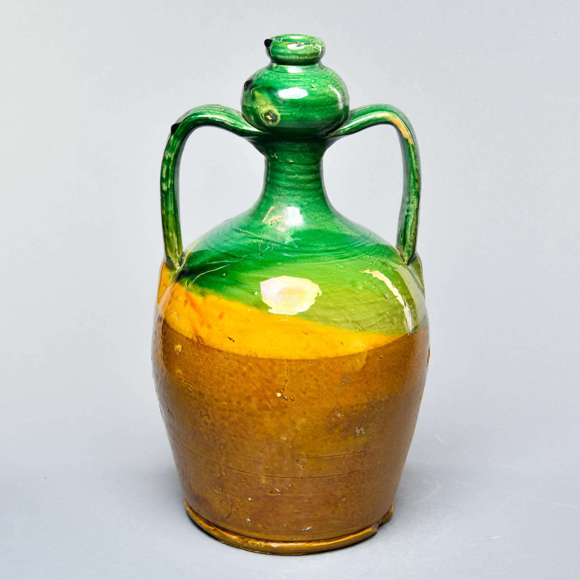 Found in France, this French vase dates from approximately 1910. This piece stands 14.75” high and has a body with 8-1/4” diameter at the base and two shoulder form handles on the sides and a narrow neck. Vessel has a dark mustard-colored glaze on