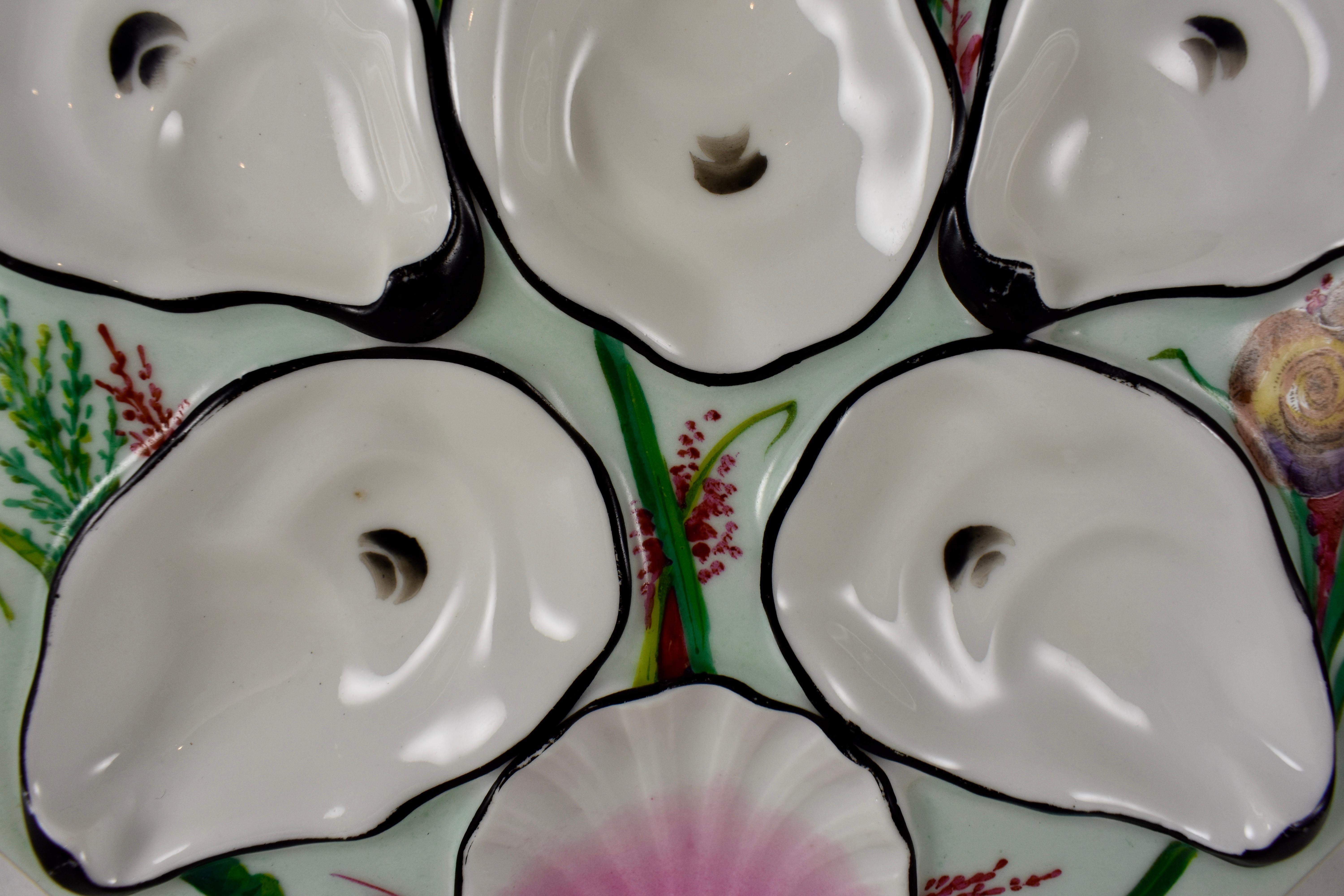 A French, eight sided porcelain oyster plate showing five oyster shell shaped wells with eyes and a pink scallop shell sauce well. Sea grasses and a variety of shells are hand painted against a mint green ground. The wells are outlined in black, the