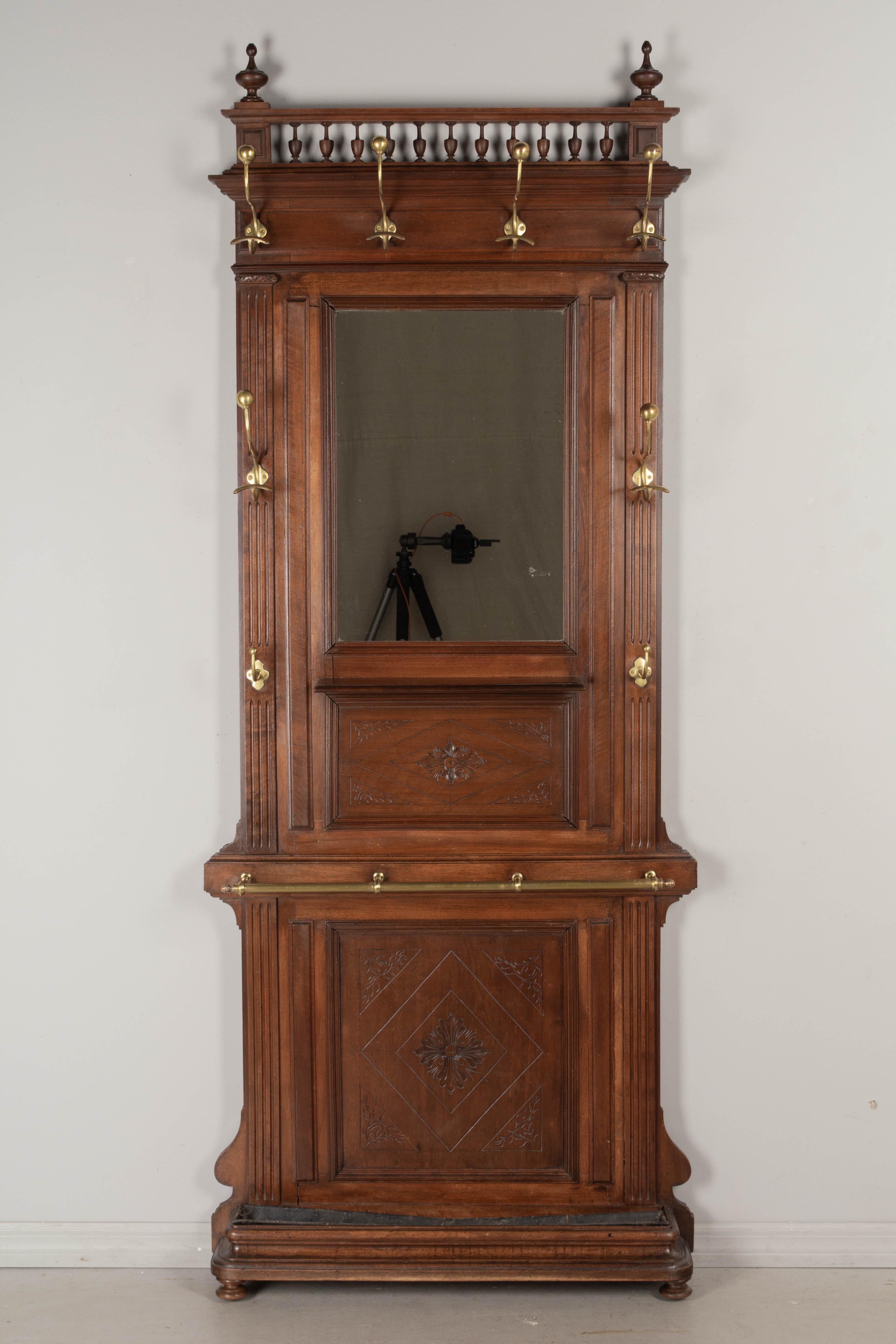 A Louis XVI style French entryway hall tree, or coat rack, with mirror. Made of solid walnut with hand carved incised decoration. Eight brass coat hooks and rail to hold umbrellas or walking canes. Small shelf in the center beneath the mirror. The