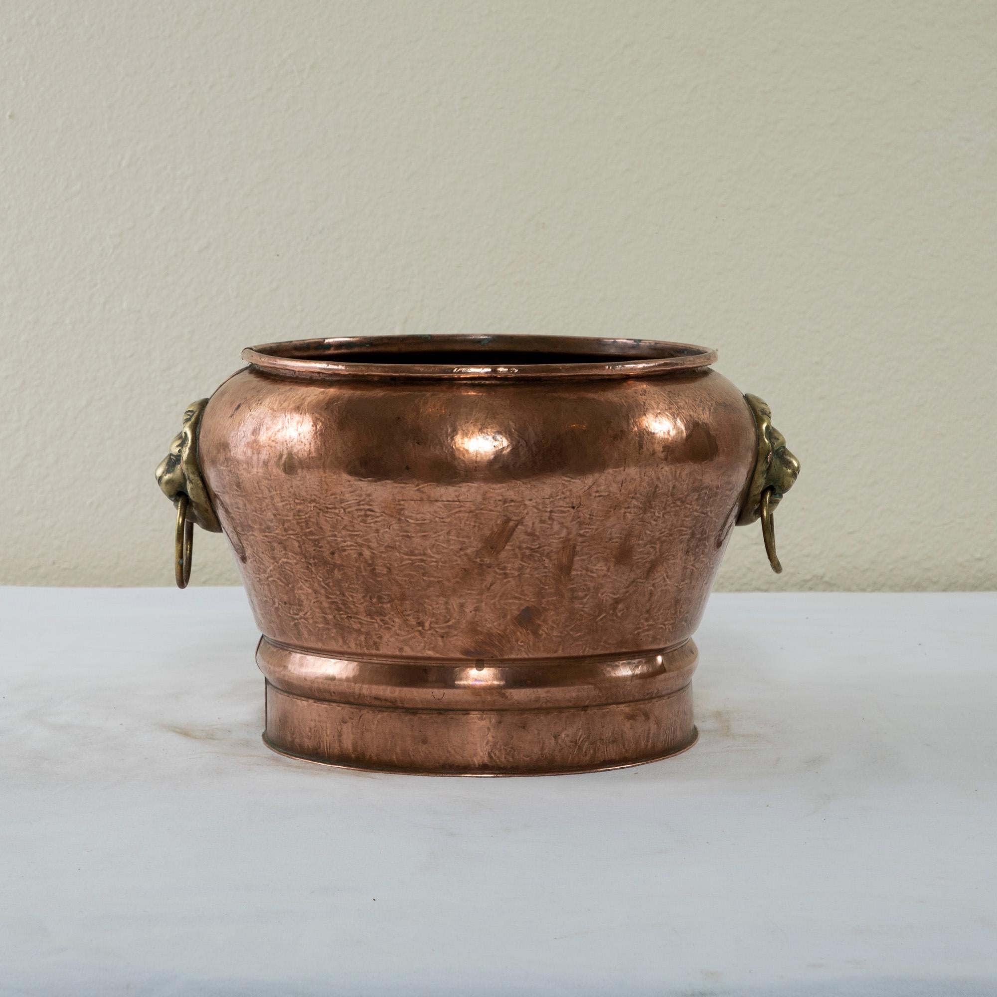 This French hand hammered copper cachepot of planter is from the turn of the twentieth century. It features a bronze lion head on each side with a drop ring handle in its mouth. The cachepot measures 8.75 inches in height by 13 inches in diameter,