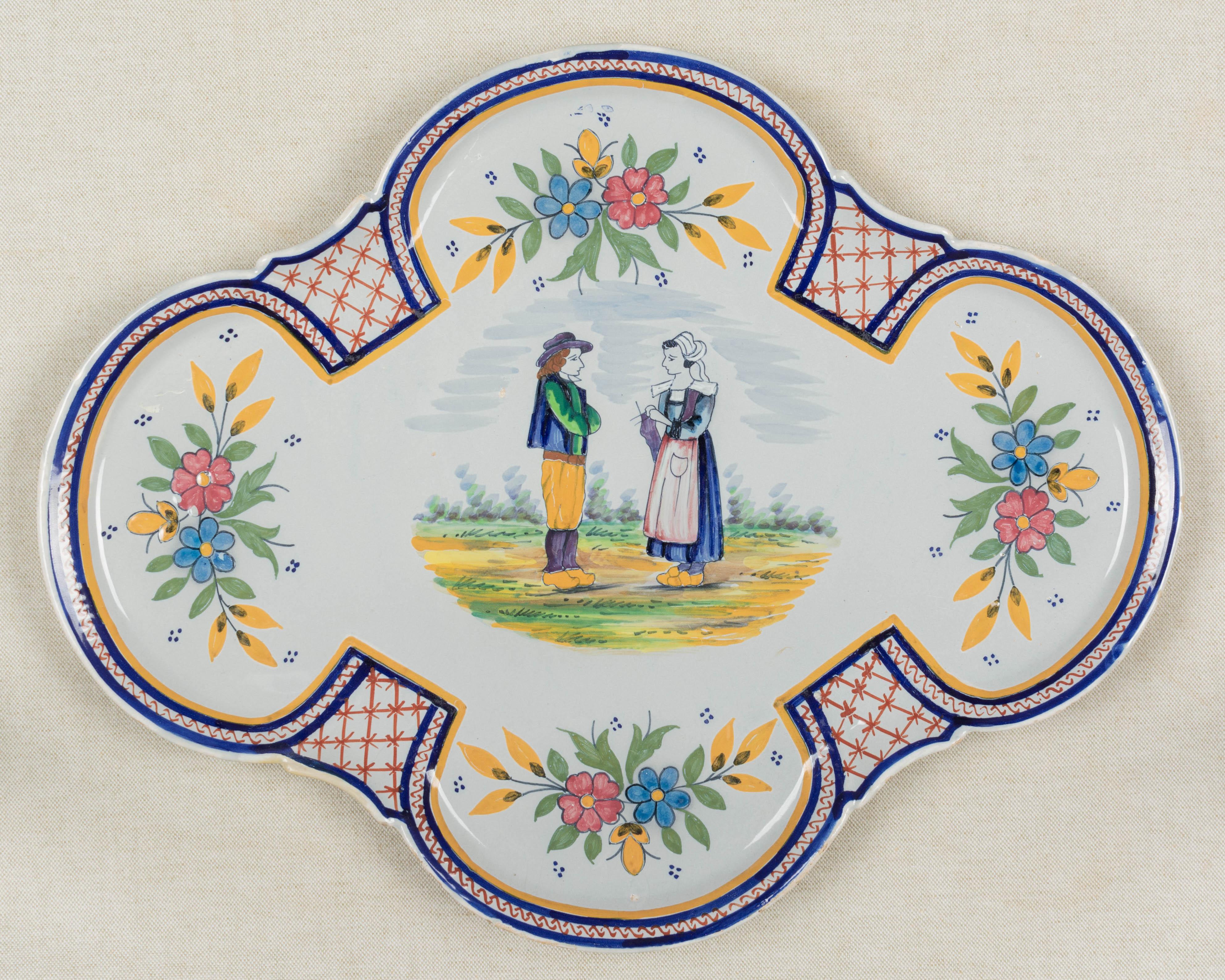Plato decorativo de loza francesa con inusual forma ovalada cuatrifoliar, pintado a mano con una pareja en el centro y decoración floral. Marca verso: HB Quimper. Reparaciones en los bordes y algunos puntos de vidriado perdido. Alrededor de
