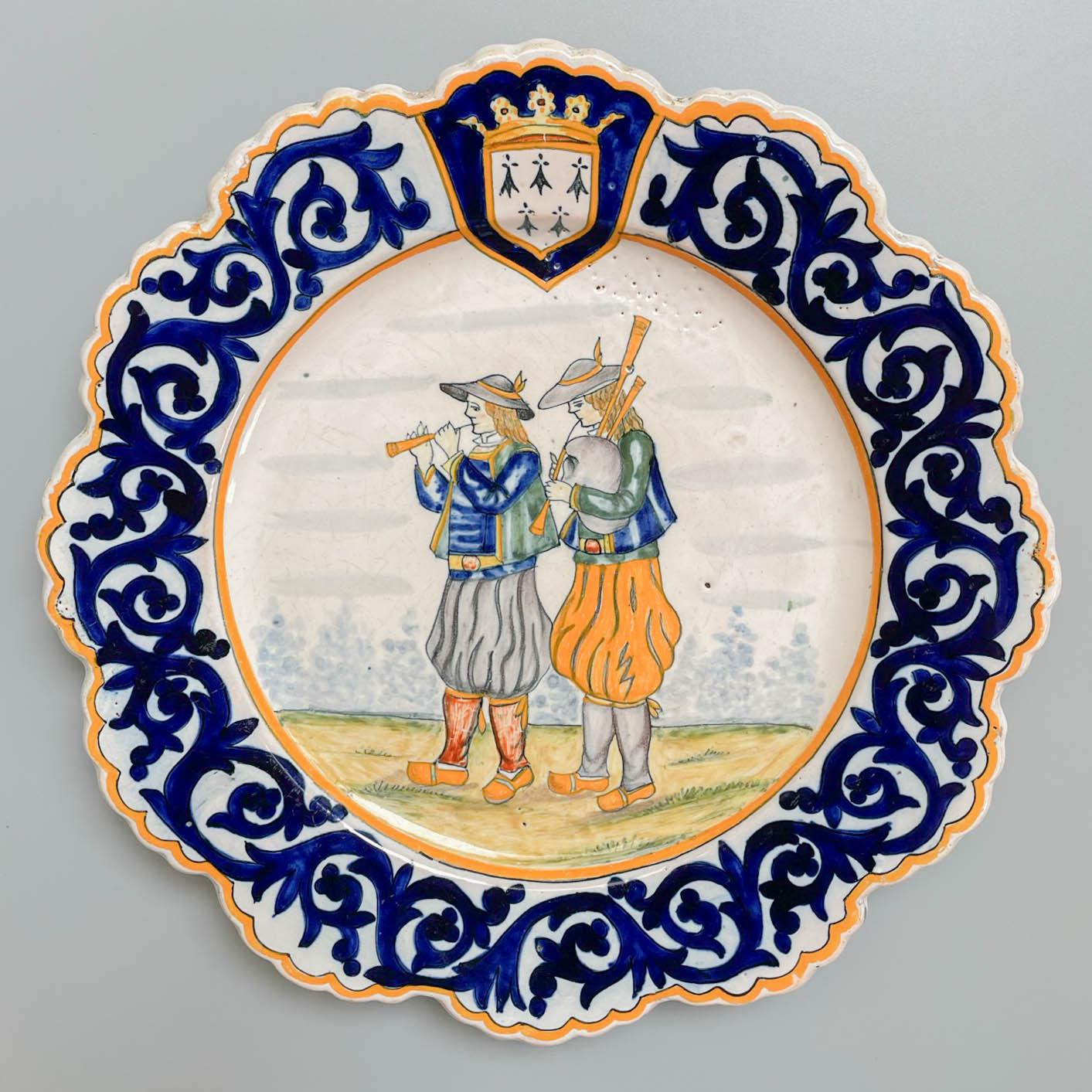 Henriot Quimper, französischer Fayence-Teller mit handgemalter Dekoration, der ein Musikerpaar darstellt, einen mit einer Pfeife und einen mit einem traditionellen Dudelsack. Blaue Blattbordüre mit bretonischem Wappen am oberen Rand. Eine gute