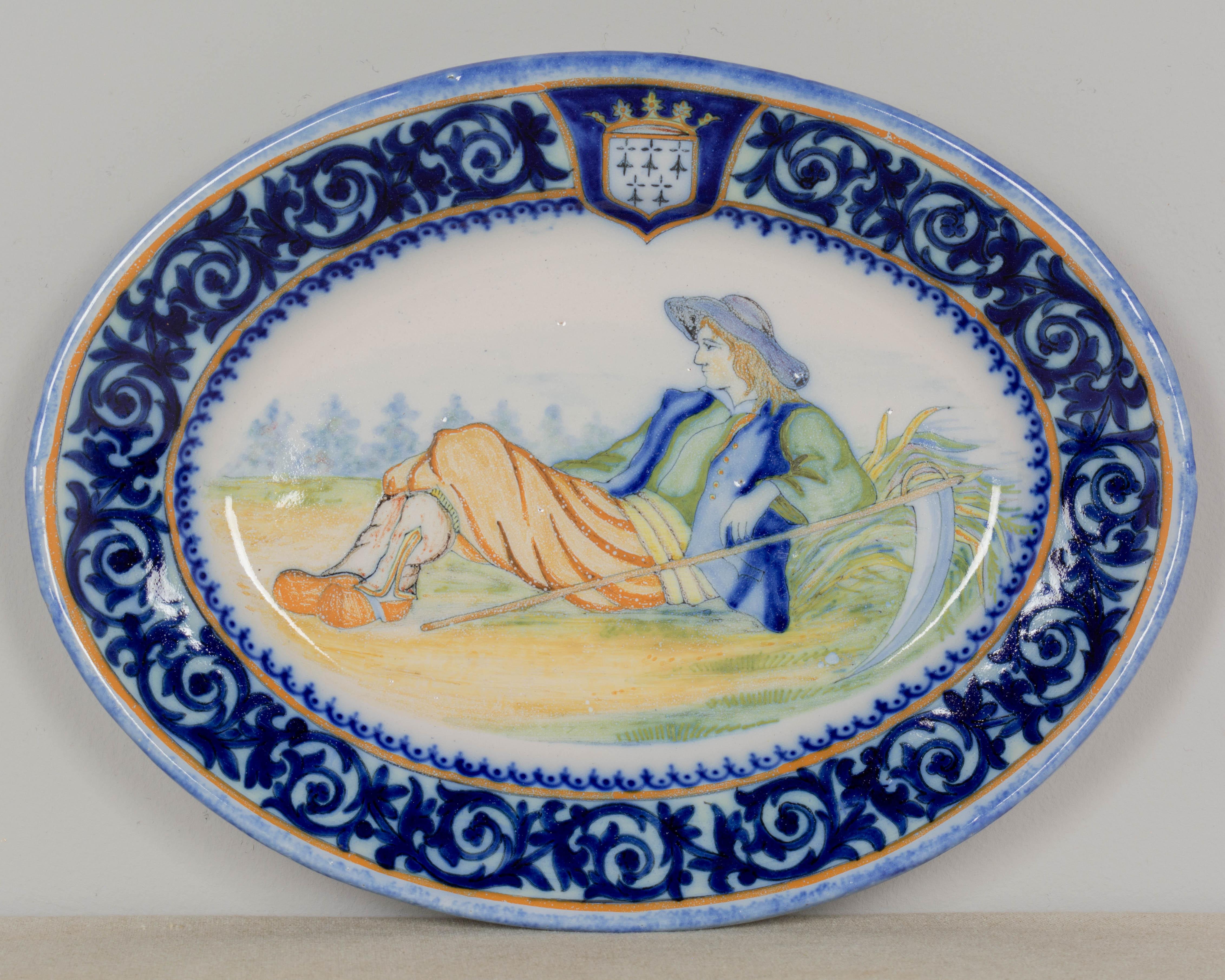 Plato ovalado decorativo de loza francesa Henriot Quimper de principios del siglo XX, escena pintada a mano de un labrador descansando con su guadaña. Amplia orla foliada azul con el escudo de Bretaña. Anverso de la marca: HR Quimper. Cerámica