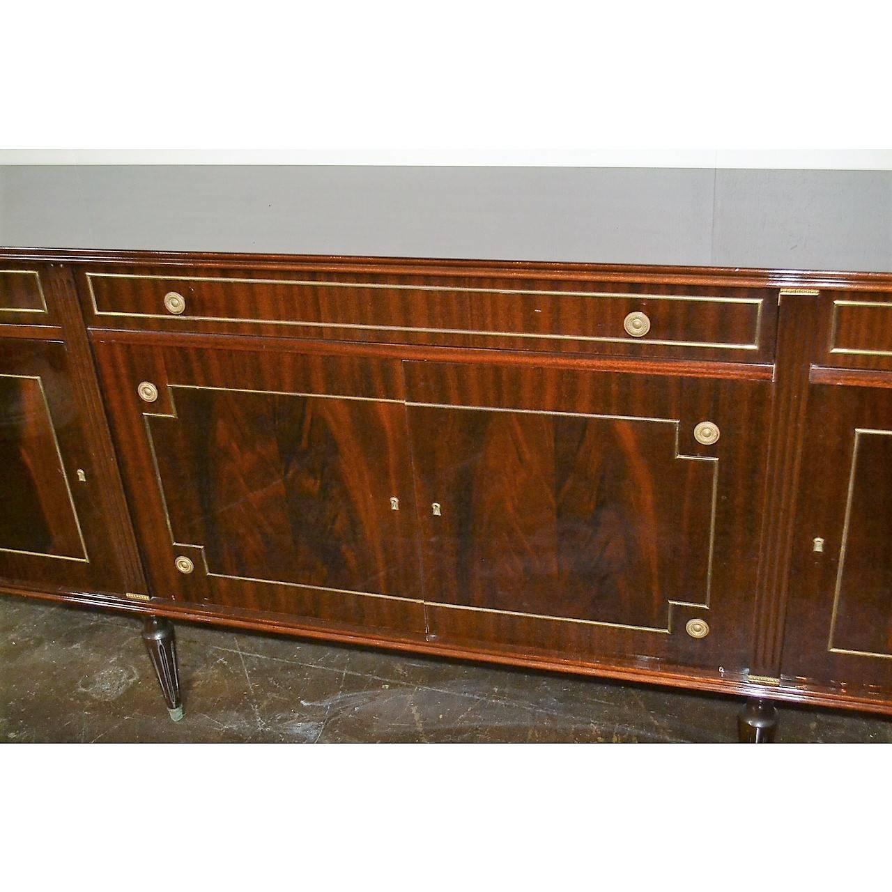 Outstanding quality and large French Jansen mahogany sideboard with superb high polish patina. The corners with fluted half columns flanking three drawers and four cupboard doors having brass accents and pulls. The whole resting on classic Louis XVI