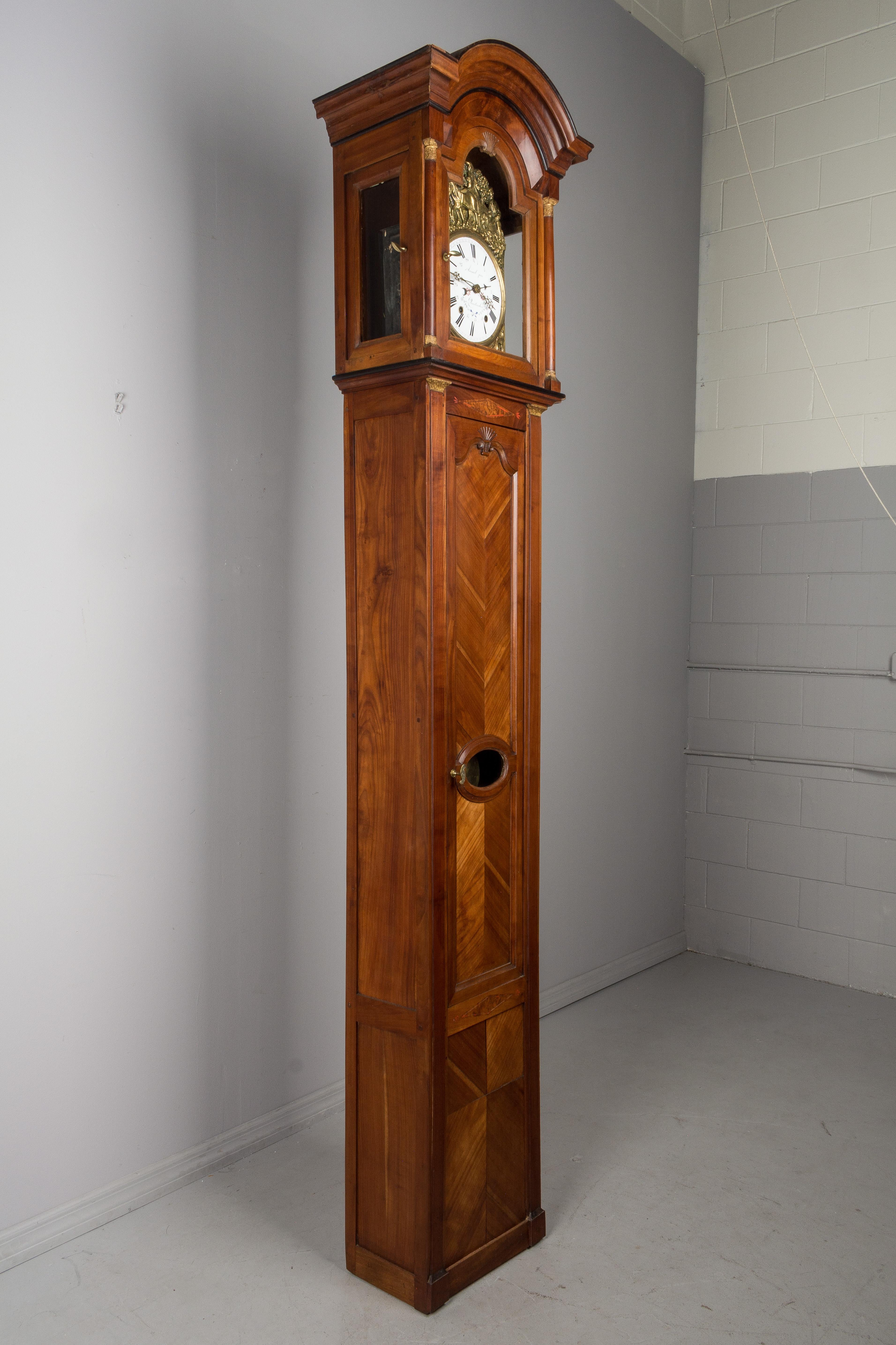 A 19th century French Horloge de Parquet or tall case clock from Brittany. Elegant solid cherry case in two parts, with book matched panels and a chapeau de gendarme crown. Subtle details including marquetry inlay and carved shell motif. Embossed