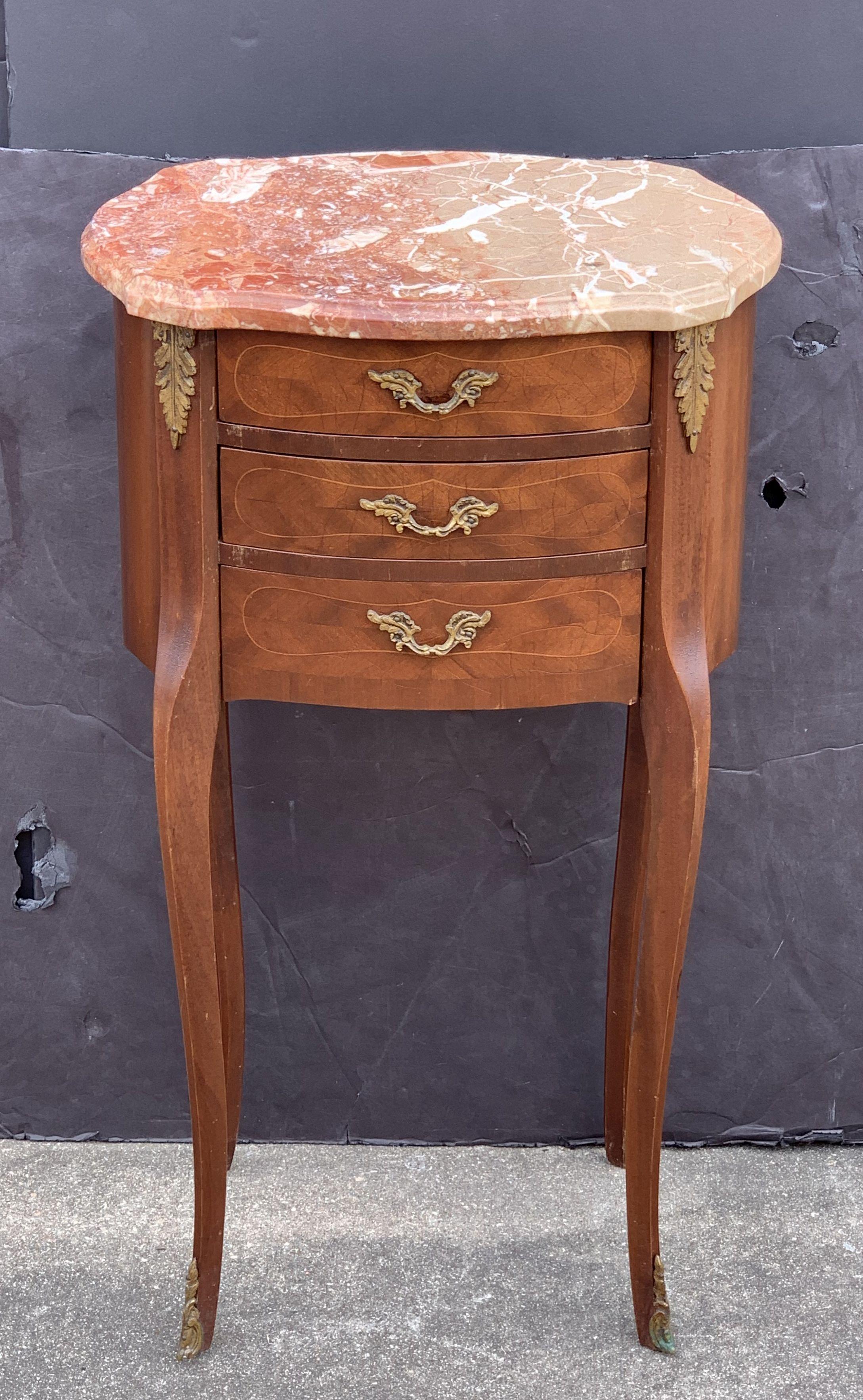 A fine pair of French bedside end tables or nightstands (or night stands) - each stand or side table featuring a figured marble-top with serpentine edge over a frieze with three inlaid drawers with decorative brass pulls, ormolu accents, and set