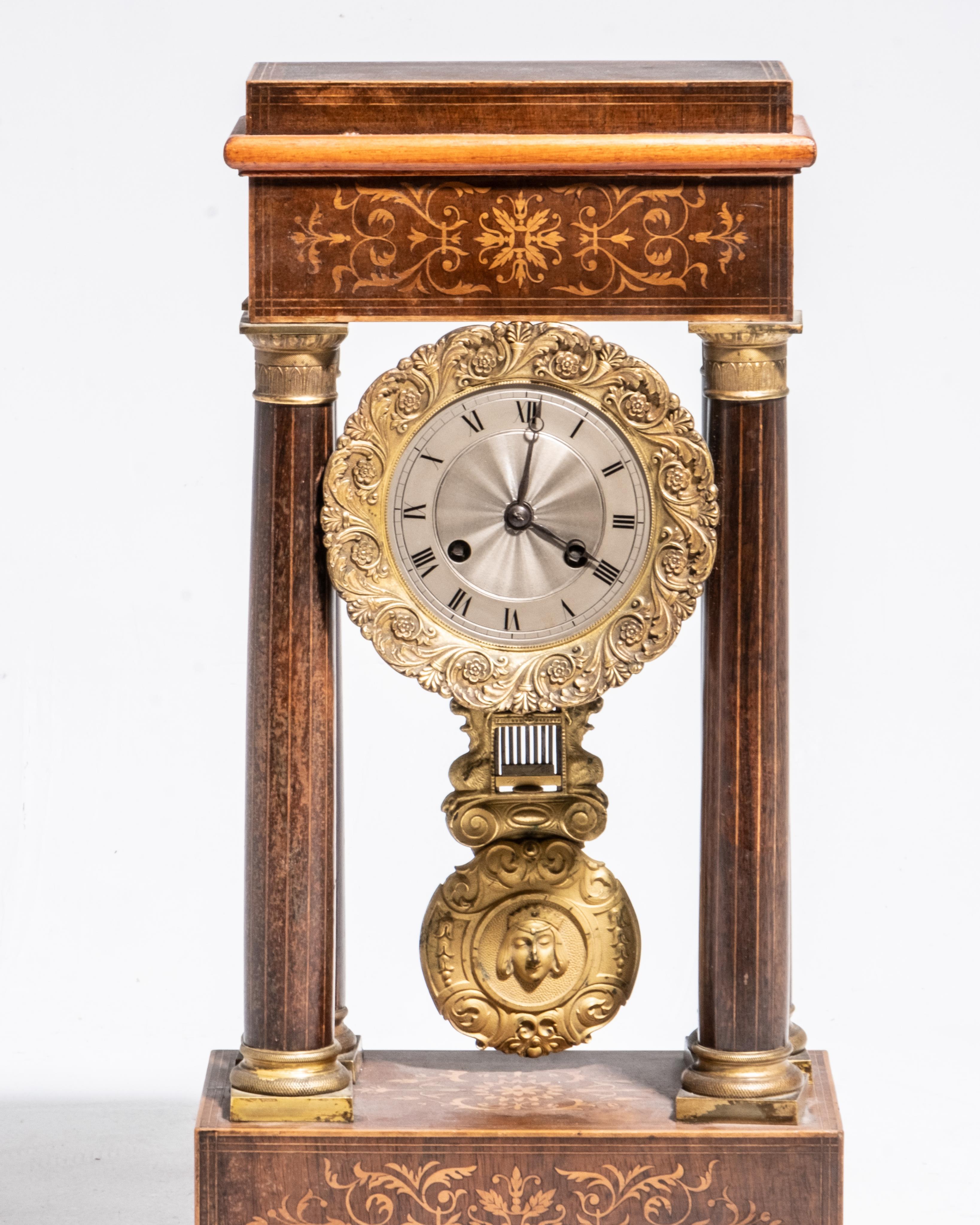French 19th-century mantle clock features a bronze and inlaid veined wood veneer portico design. The clock is adorned with gilded ornaments, including a decorative pendulum and intricate floral inlays on the base and top. The dial, is surrounded by