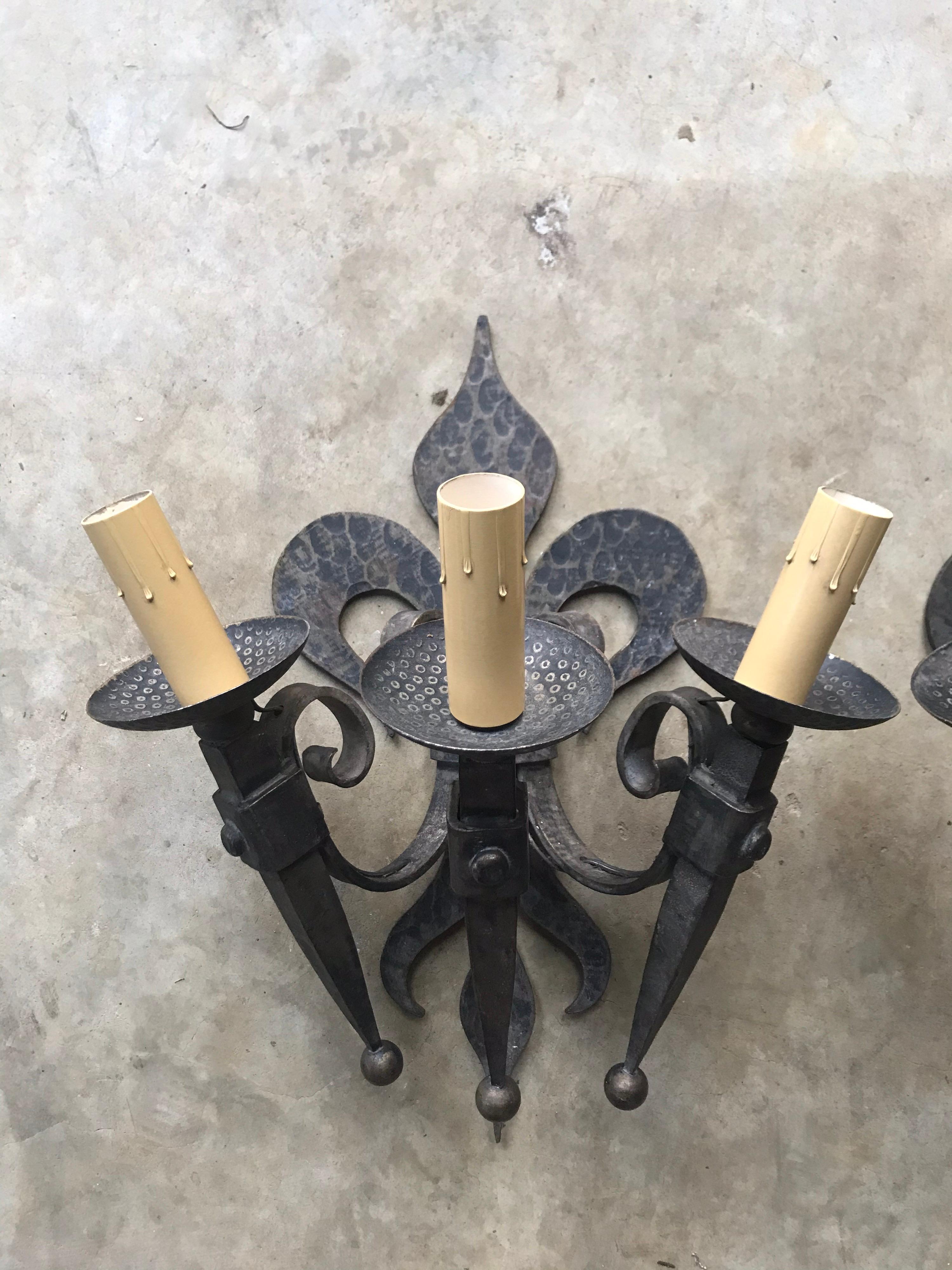 Pair of French iron 3-arm sconces. Made of heavy wrought iron, these impressive sconces feature a hammered fleur-de-lys back with 3 attached arms. These sconces have a gothic/chateau feel to them and provide lots of light with 40 watt bulbs for each