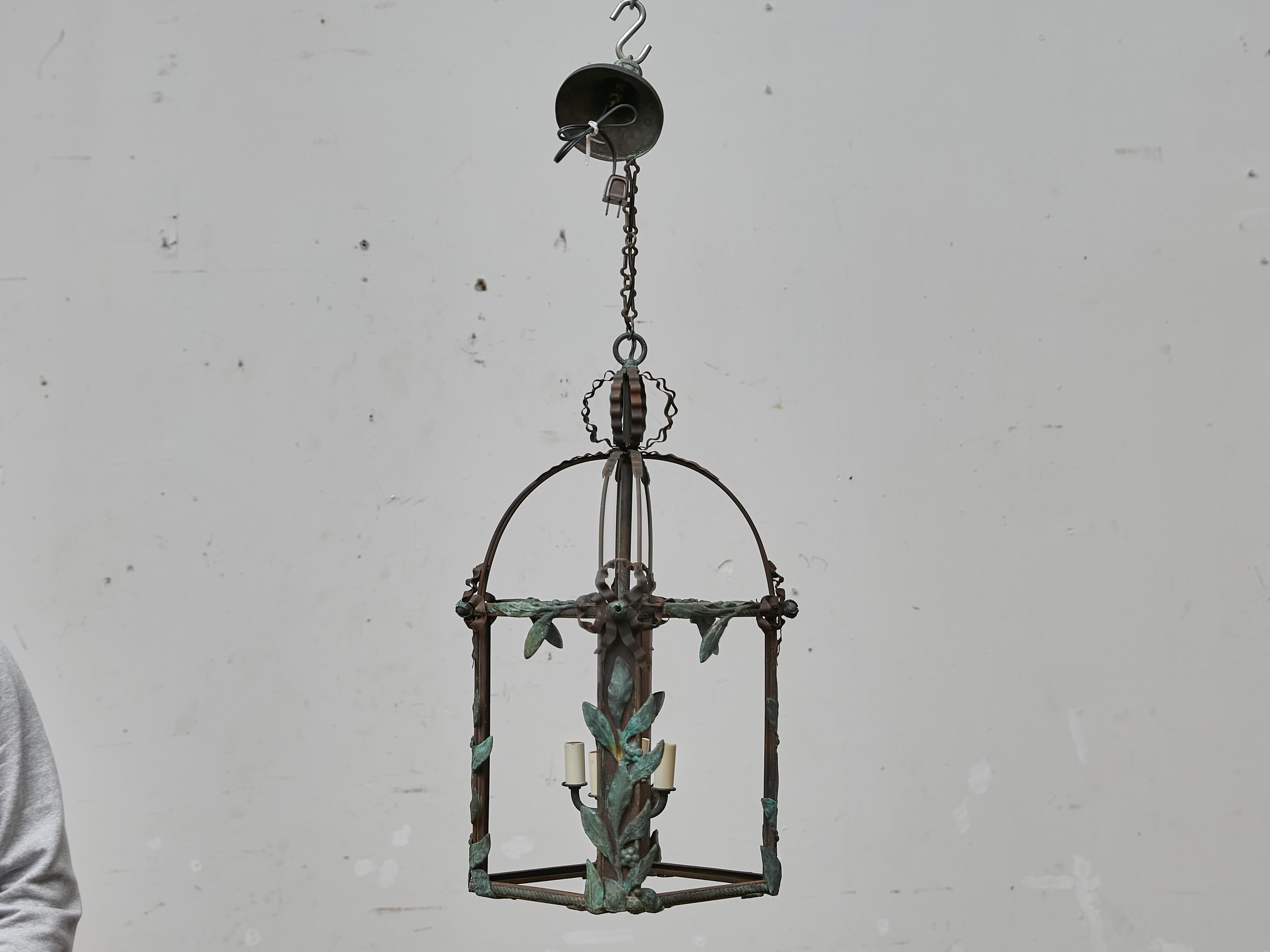Questa incantevole lanterna francese del 1920 circa è caratterizzata da un gioco scultoreo di ferro battuto e rame patinato verdastro, che formano una delicata gabbia a cupola con accenti botanici. La struttura aperta è definita da nervature in