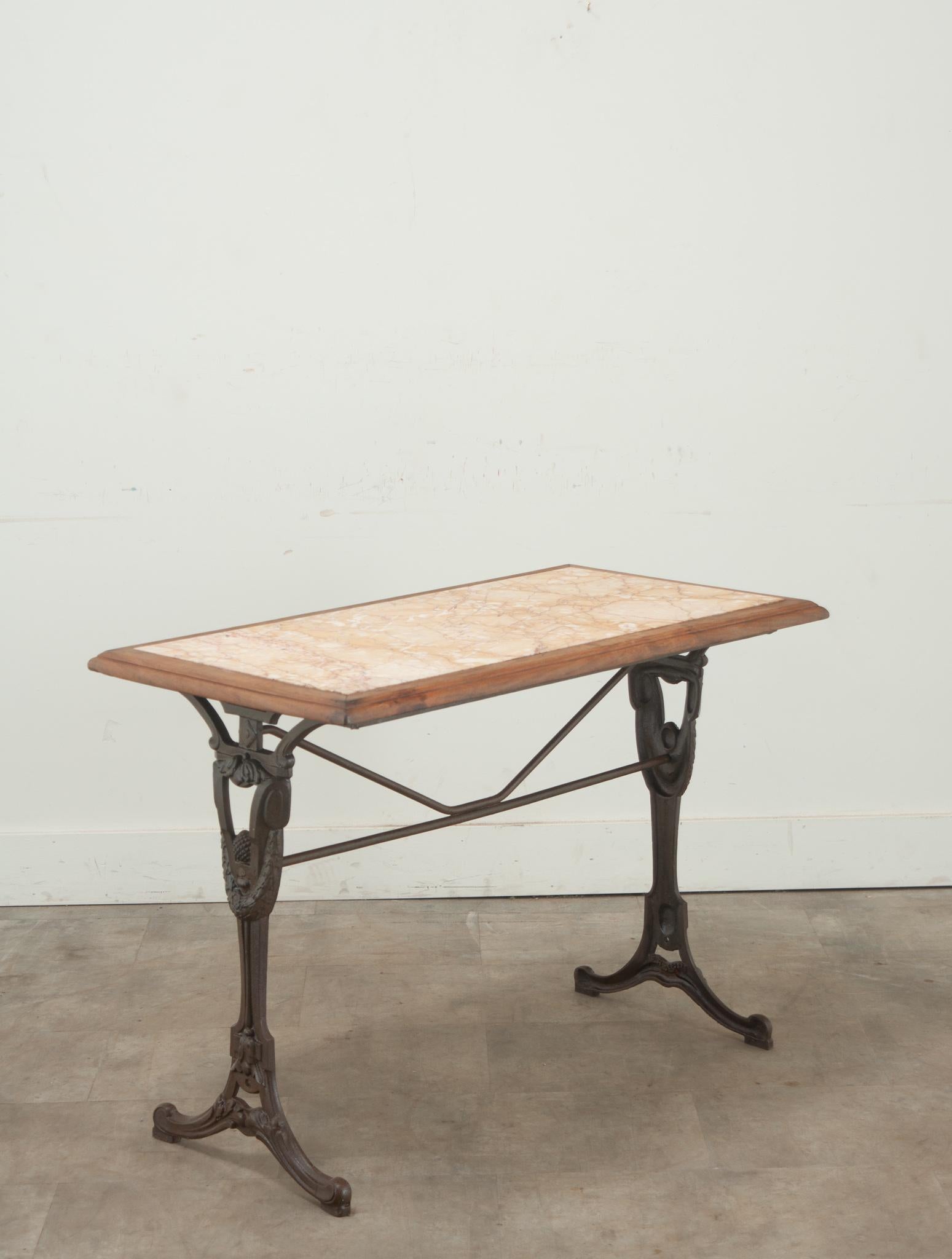 A classically styled French bistro table featuring a uniquely colored marble surrounded in a walnut frame. The top sits over a cast iron frame connected by a stretcher for additional support. There is wear consistent with age and use, be sure to