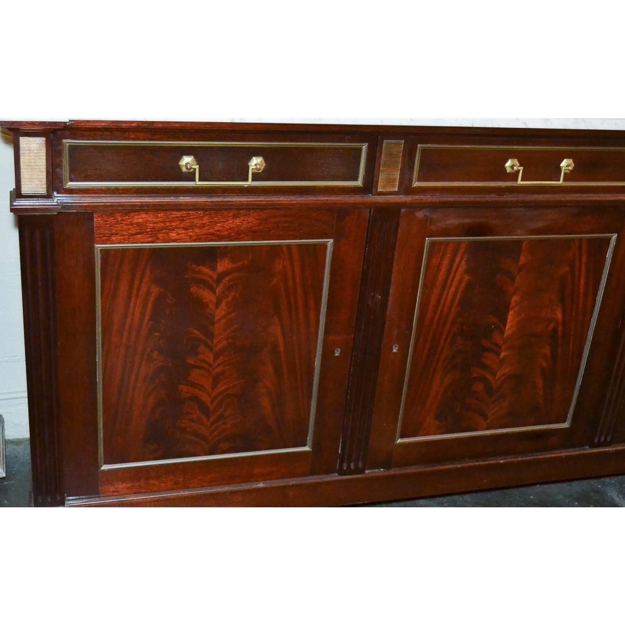 Fabulous French Jansen flame mahogany server or sideboard with a superb white Carrara marble top. The three drawer frieze with brass pulls, trim, and ribbed brass inlays above three cupboard doors. The entire resting on turned and tapered