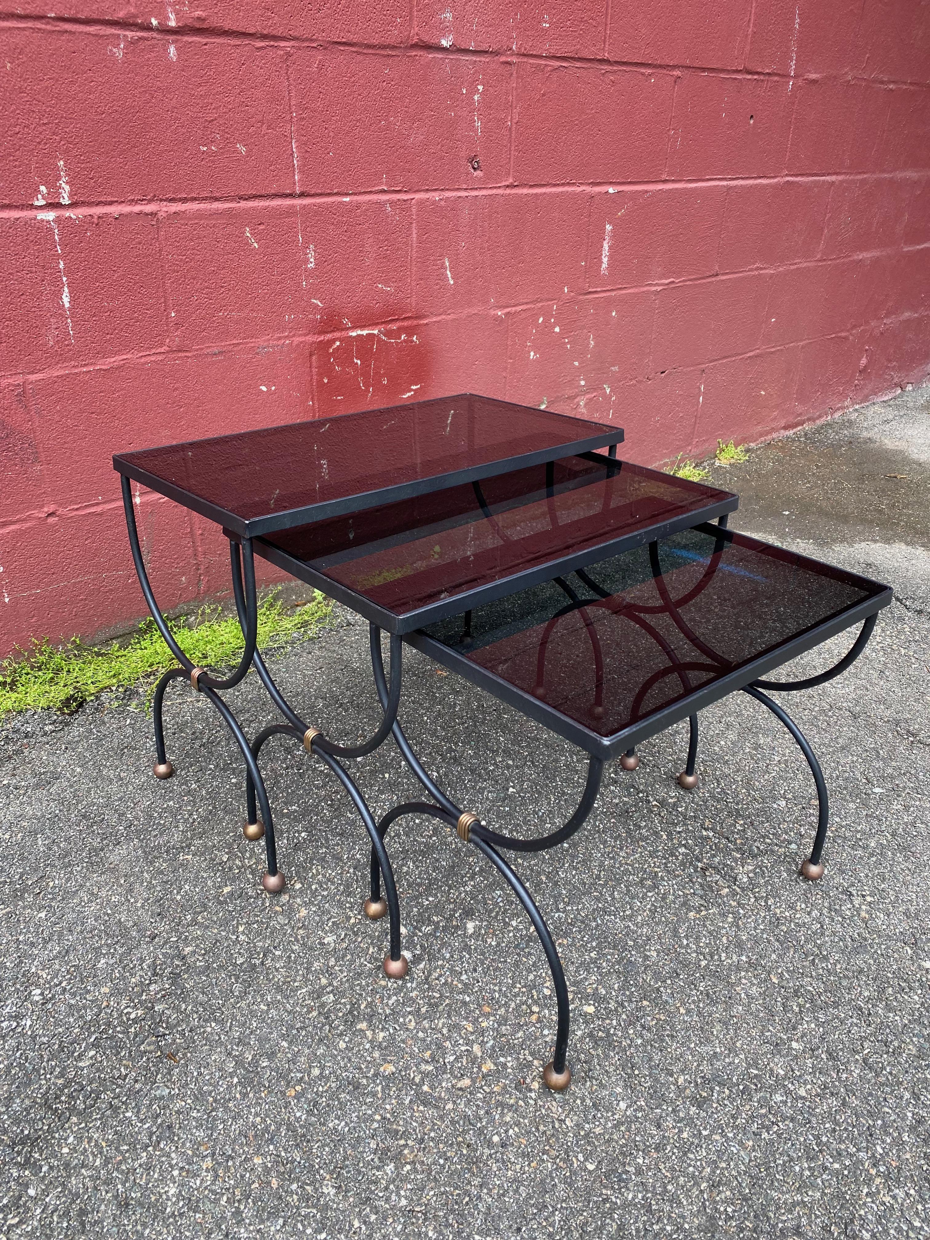 Set of three French Mid Century nesting tables with new black glass nestled in wrought iron frames. The curved black cylindrical frames feature polished brass accents and ball feet in the style of Jean Royere. Wherever they are placed, this stylish