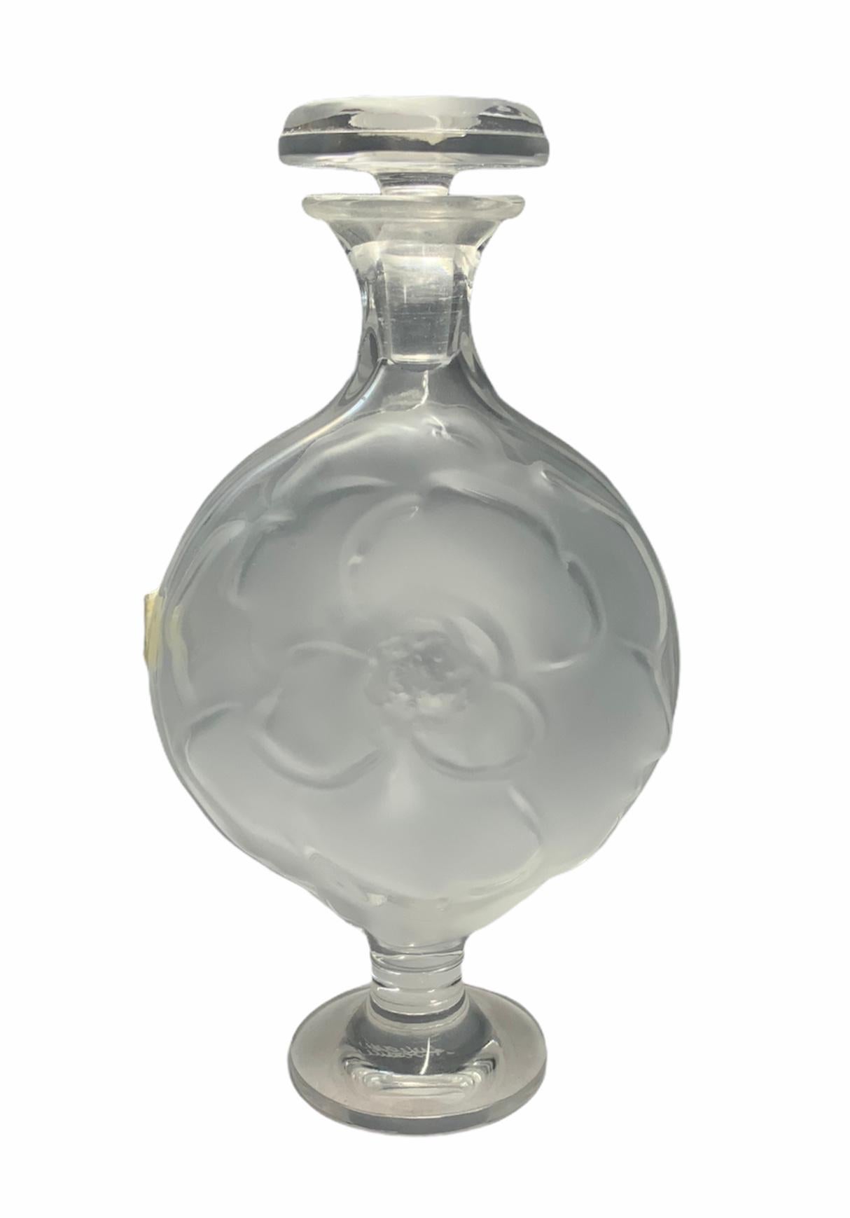 This is a Lalique glass perfume bottle shaped as a round flat urn. It is decorated with a frosted rose in one side and a camellia flower in the other side. The flat round shaped stopper is also decorated with a frosted camellia. Under the base is