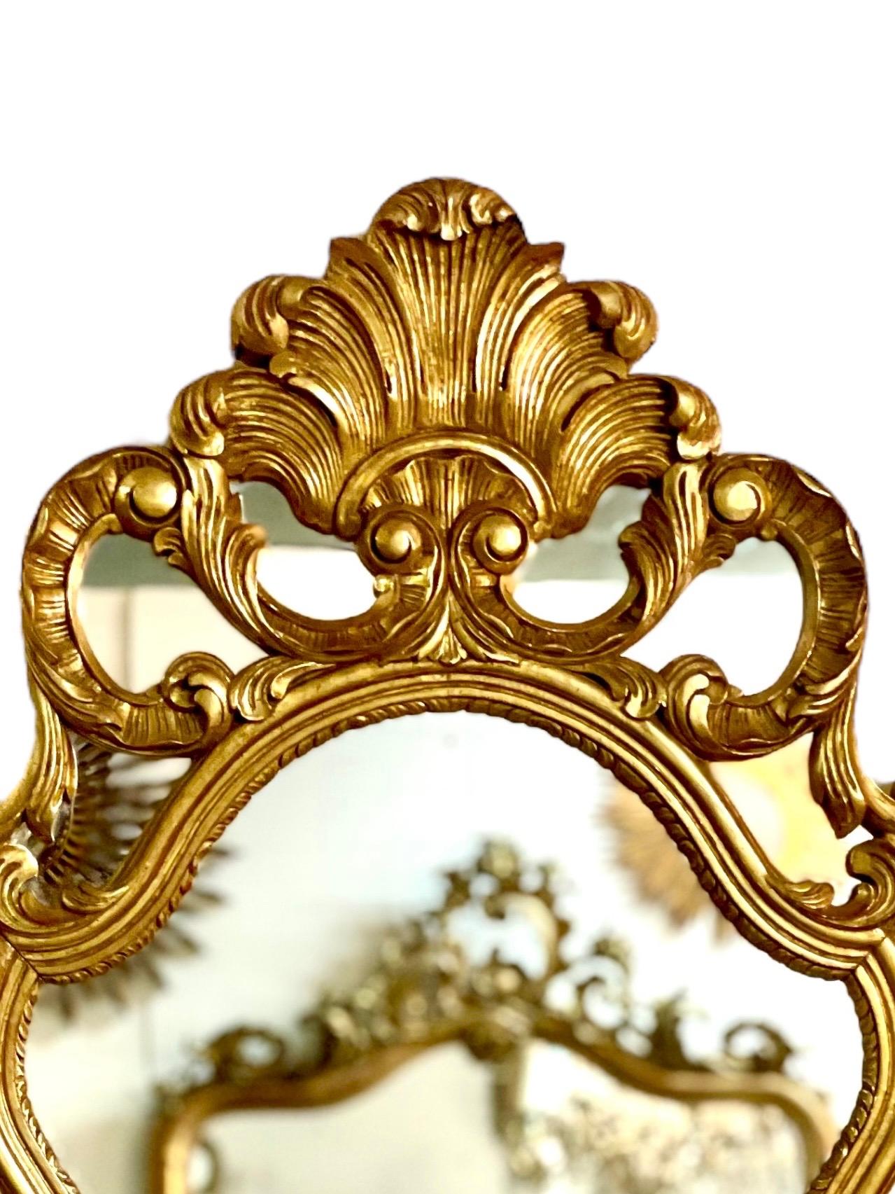 Dieser prächtige vergoldete Wandspiegel im Rokoko-Stil hat einen sehr verzierten Rahmen, der mit Spiegelplättchen besetzt ist und mit einem kunstvollen Wappen aus stilisierten Federn gekrönt wird. Dieser große und beeindruckende Spiegel aus dem 20.