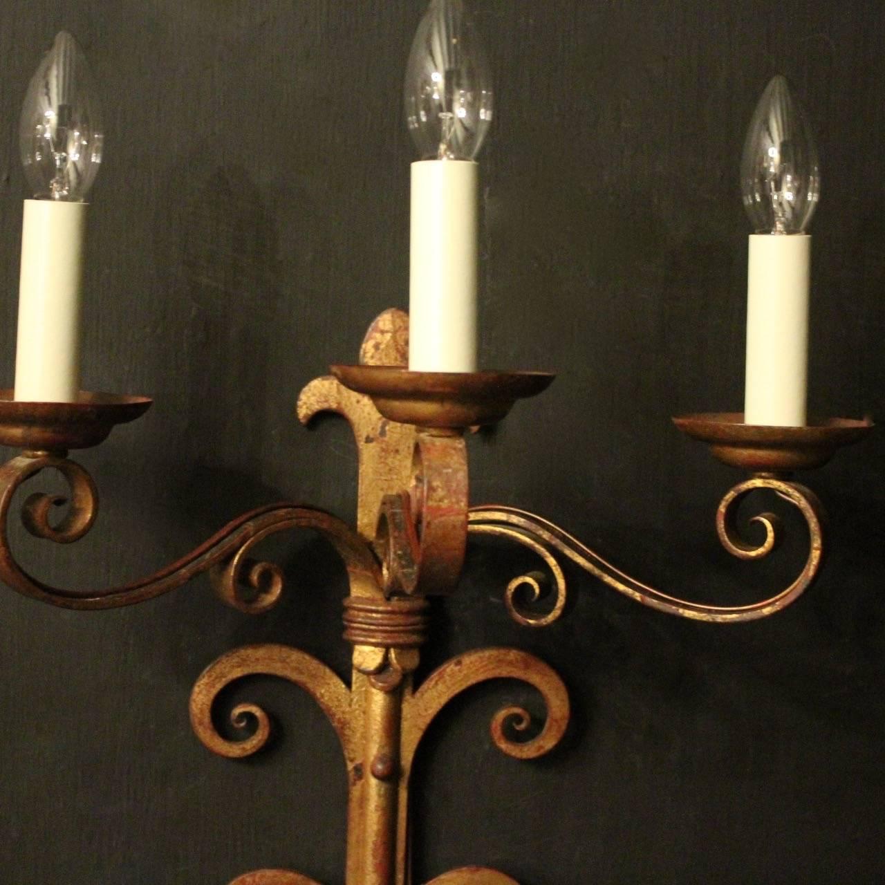 A large decorative pair of French gilded wrought iron triple arm antique wall lights, the rectangular gauge scrolling arms with circular bobeche drip pans, issuing from an elongated twin scrolled backplate with fleur de lys finial and terminal, nice