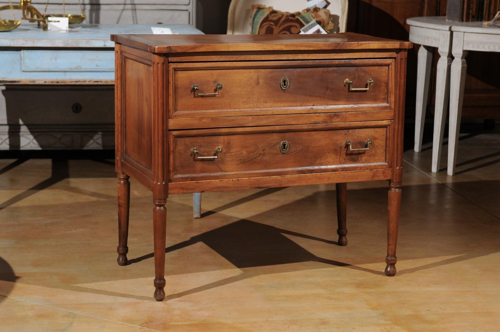 A petite French Louis XVI period walnut commode from the late 18th century, with two drawers and fluted accents. Born in France during the reign of King Louis XVI, this exquisite petite commode features a rectangular top with rounded corners,