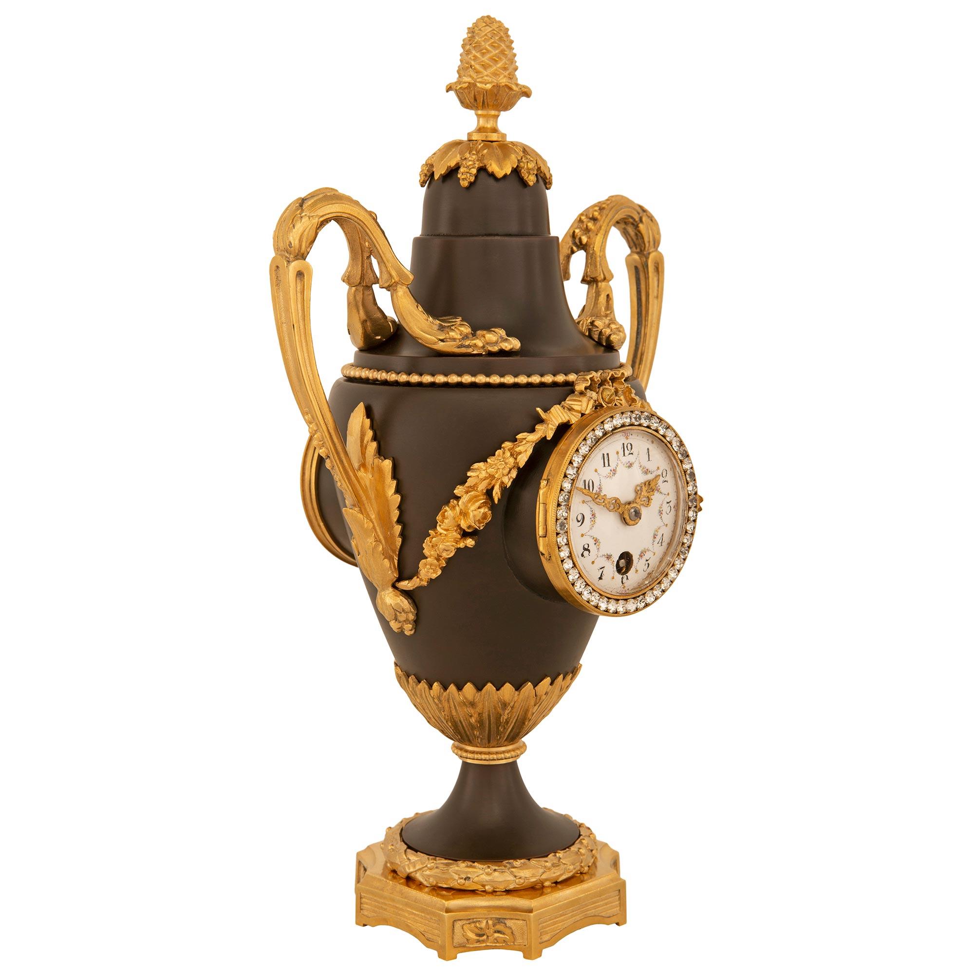 Elégante pendule en bronze patiné et bronze doré de style Louis XVI de la fin du XIXe siècle. L'horloge en forme d'urne de prospérité est surélevée par une base carrée aux angles concaves et aux panneaux en retrait. Les panneaux concaves sont