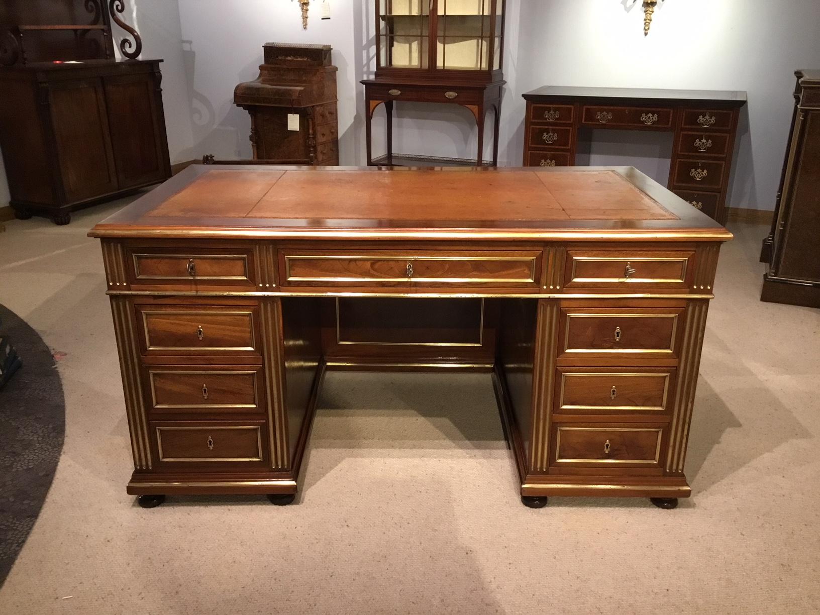 A French late 19th century mahogany and brass-mounted antique writing desk. The rectangular solid mahogany top having brass moulding, a tan leather inset writing surface with gilt tooled detail and twin secretary slides. Having an arrangement of