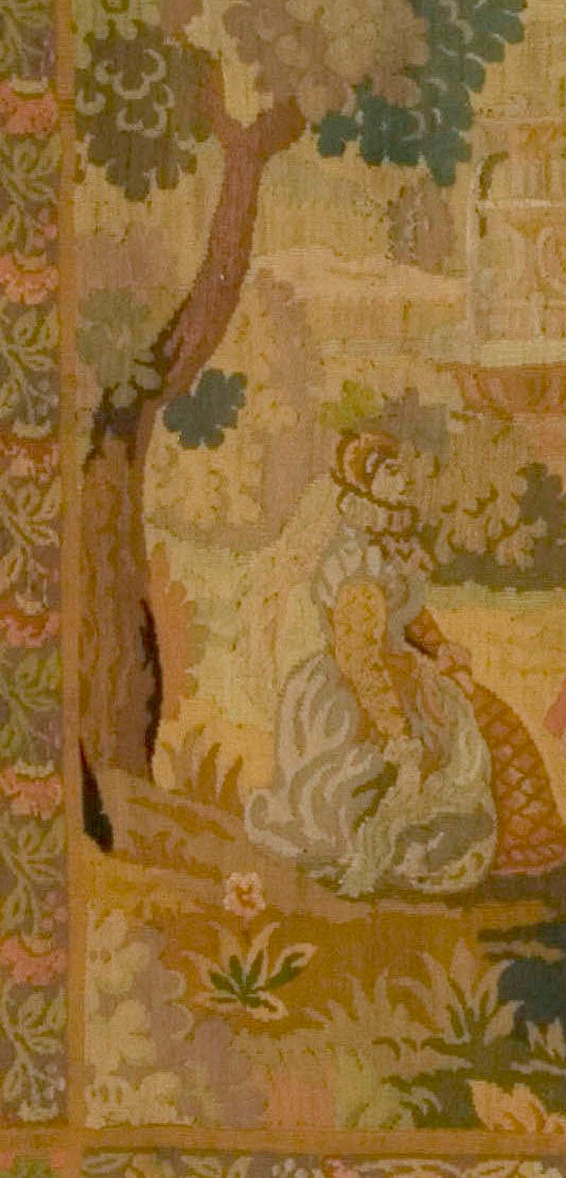 A French Hand Loomed circa 1890 tapestry. A noblewomen being amused by a servant playing a lute in a formal garden setting in the background is a stately home and the noblewomen is siting under a tee for shade, all surrounded by a floral border. 