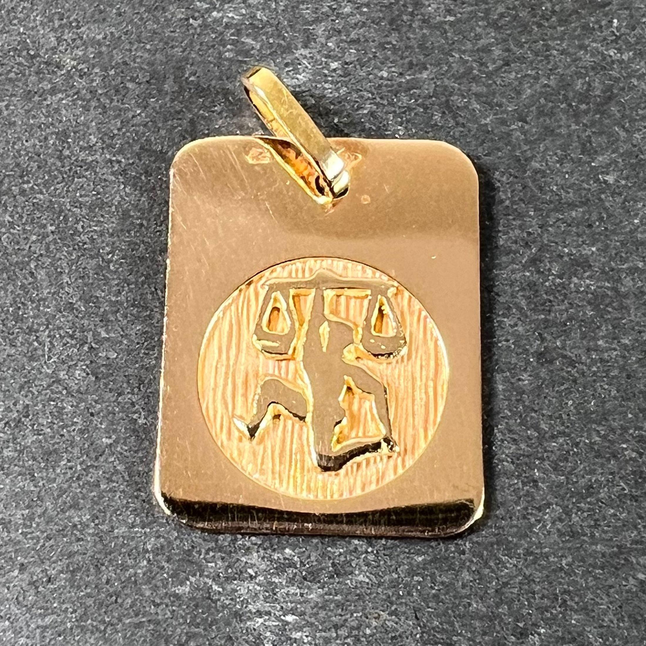 Ciondolo charme francese in oro giallo 18 carati (18K) disegnato come una medaglia rettangolare con un incavo circolare inciso che raffigura un uomo che solleva una bilancia sopra la sua testa e rappresenta il segno zodiacale della Bilancia. Timbro
