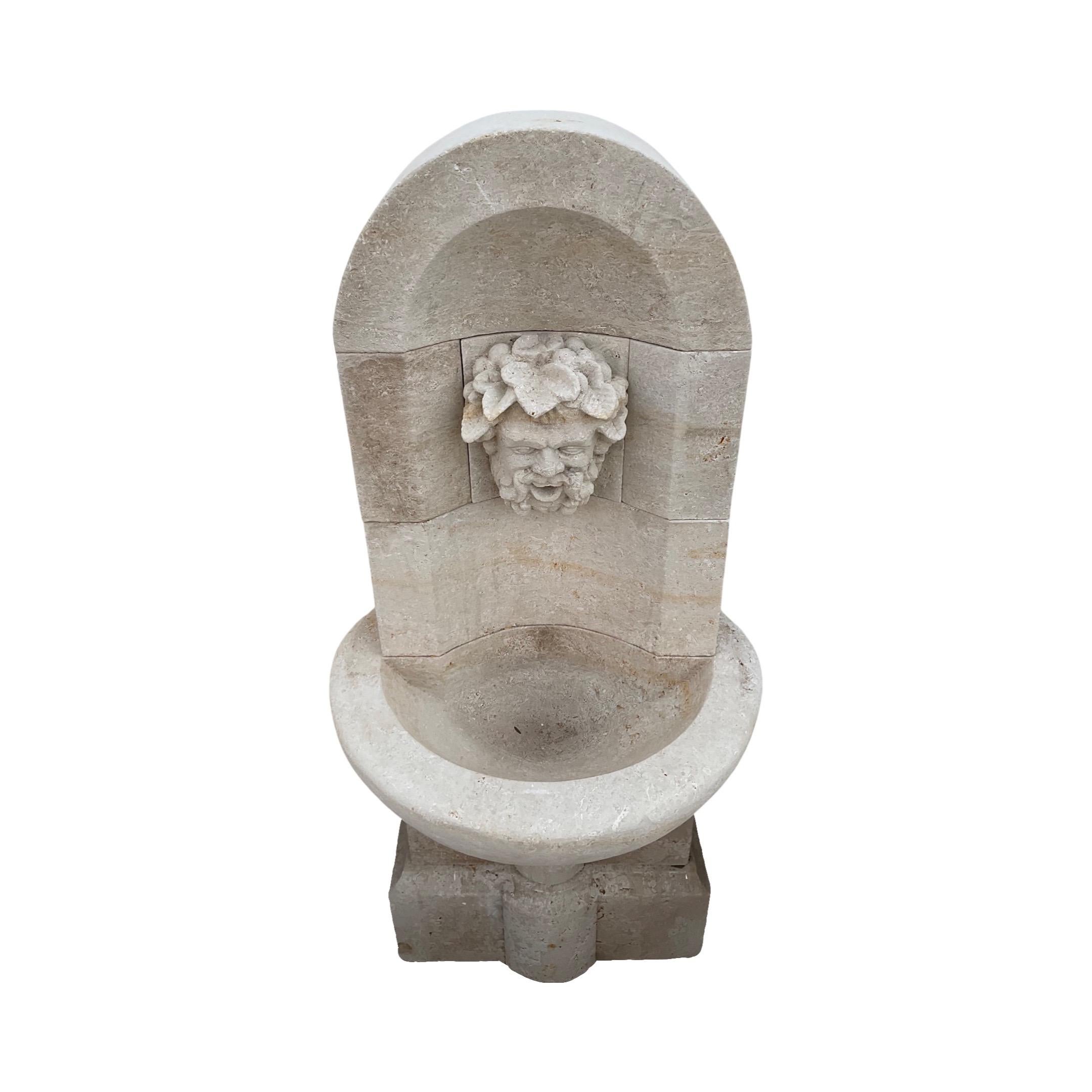 Beautify your outdoor settings with this small, mid-century French Limestone Wall Fountain. Crafted from limestone, this Louis XVI-style fountain features a carved face in the center, serving as a water exit, along with a small basin for water