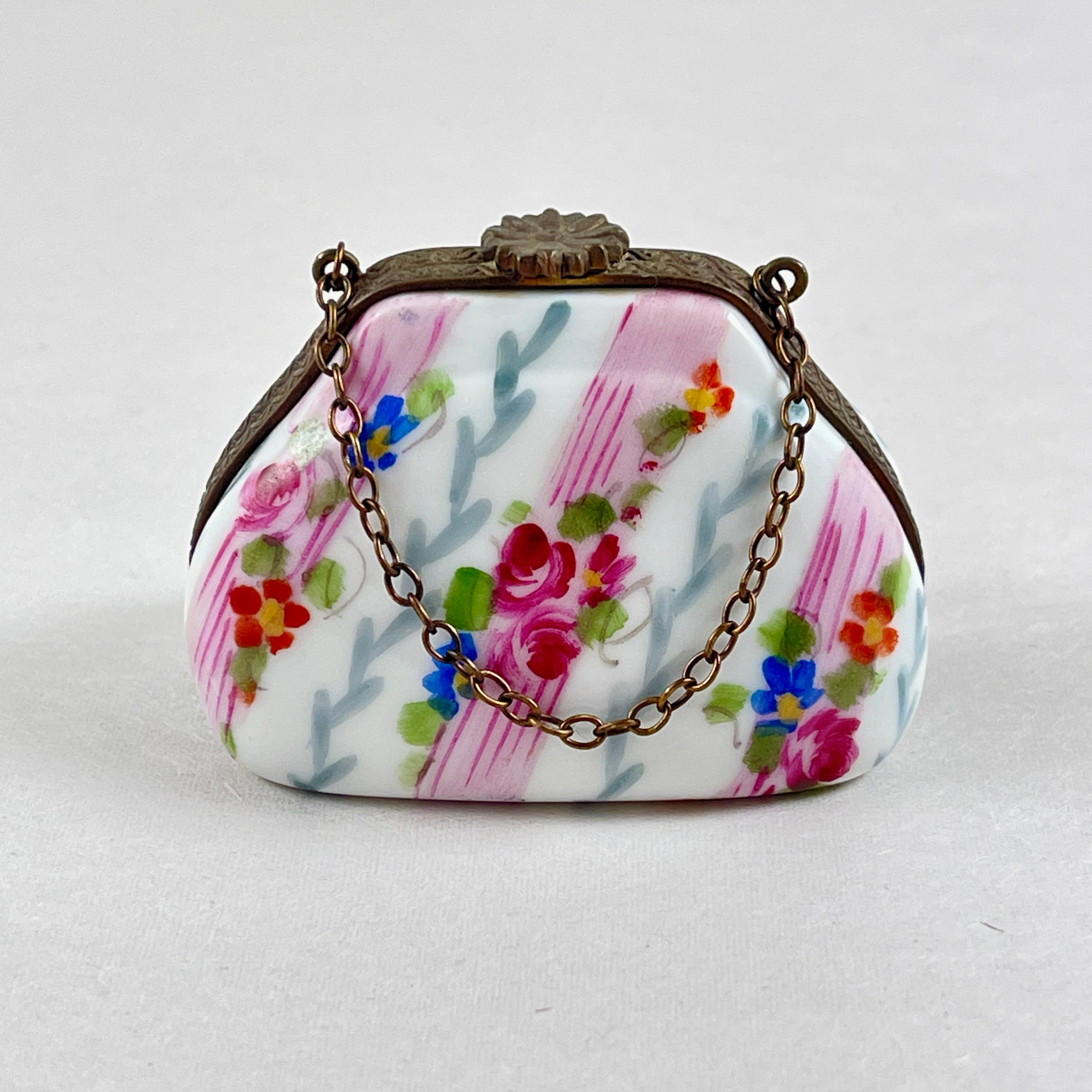 Caja de baratijas francesa de Limoges pintada a mano en forma de bolso con asa de cadena, hacia la década de 1980.

Un encantador motivo floral de rosas y rayas rosas, reforzado en metal dorado con un broche floral.

Se abre para mostrar dos plumas