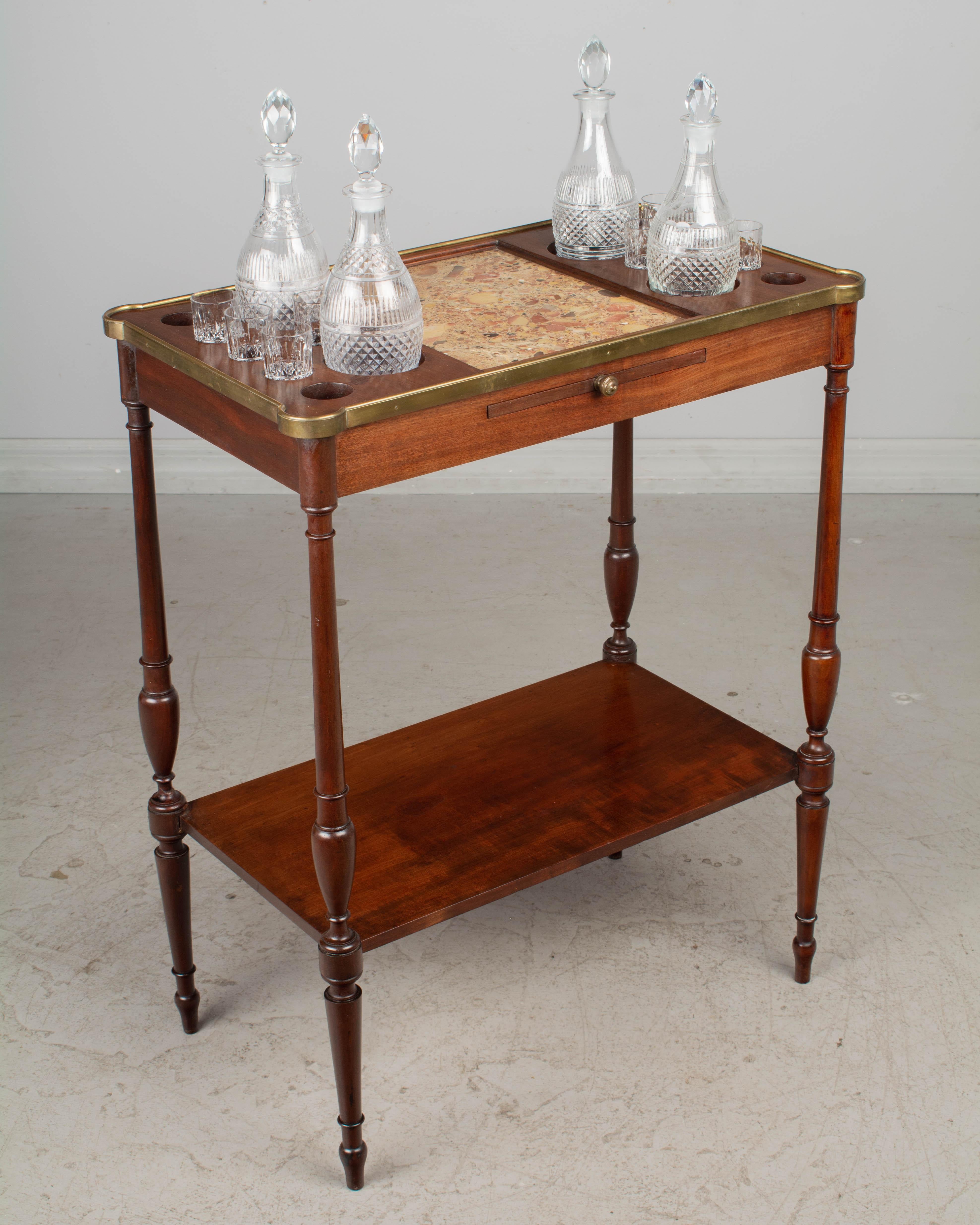 A French Louis XVI style liquor side table, or small bar, made of solid mahogany with lower shelf, turned tapered legs and a pull out board for serving. The top has a brass surround, a Breche d'Alep marble inset and wells to hold bottles and