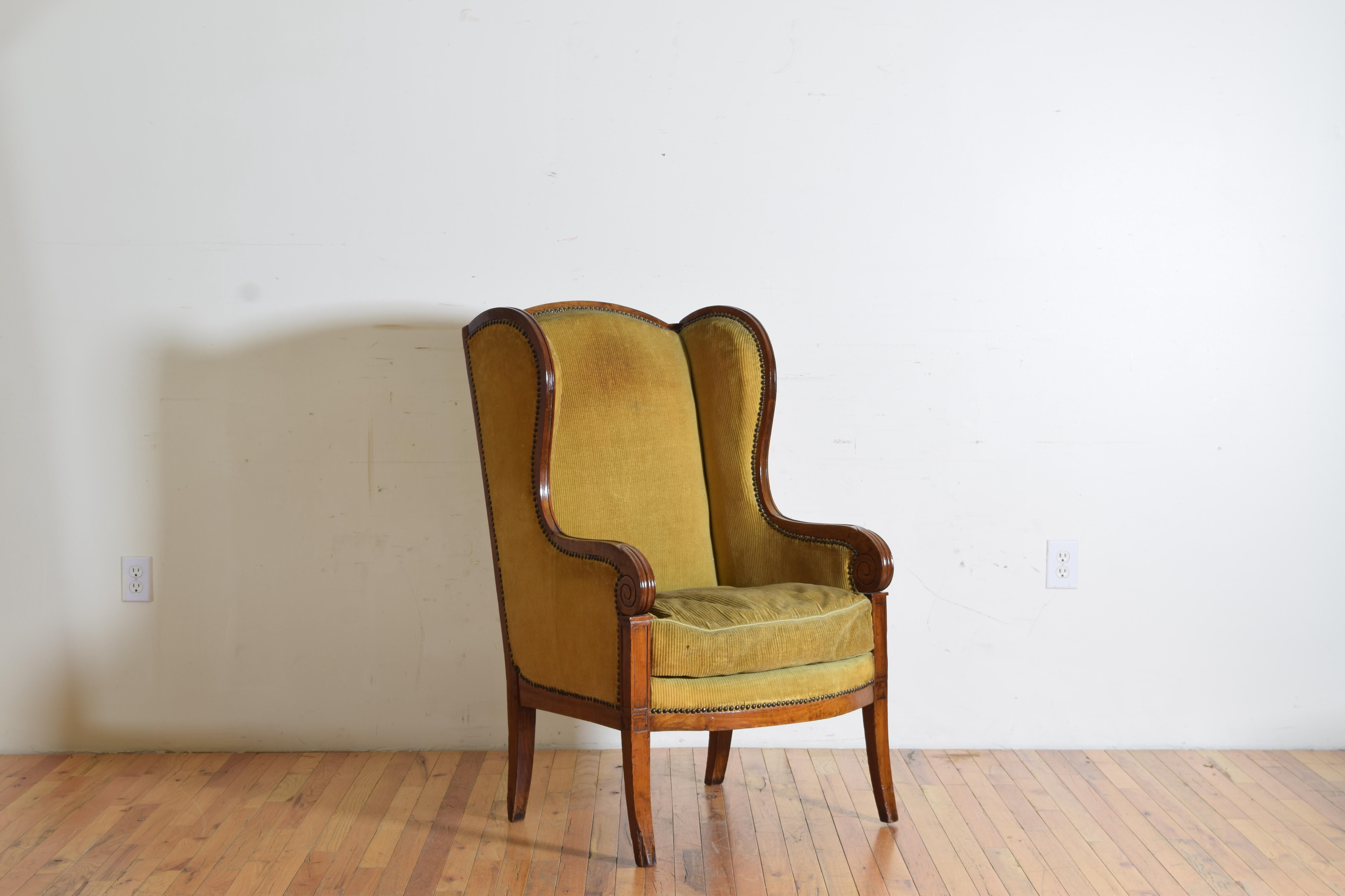 Having an arched backrest flanked by arched and shaped wings descending into carved handles, the loose cushion above a fitted deck and raised on flared tapering legs, the vintage corduroy upholstery in very good condition.