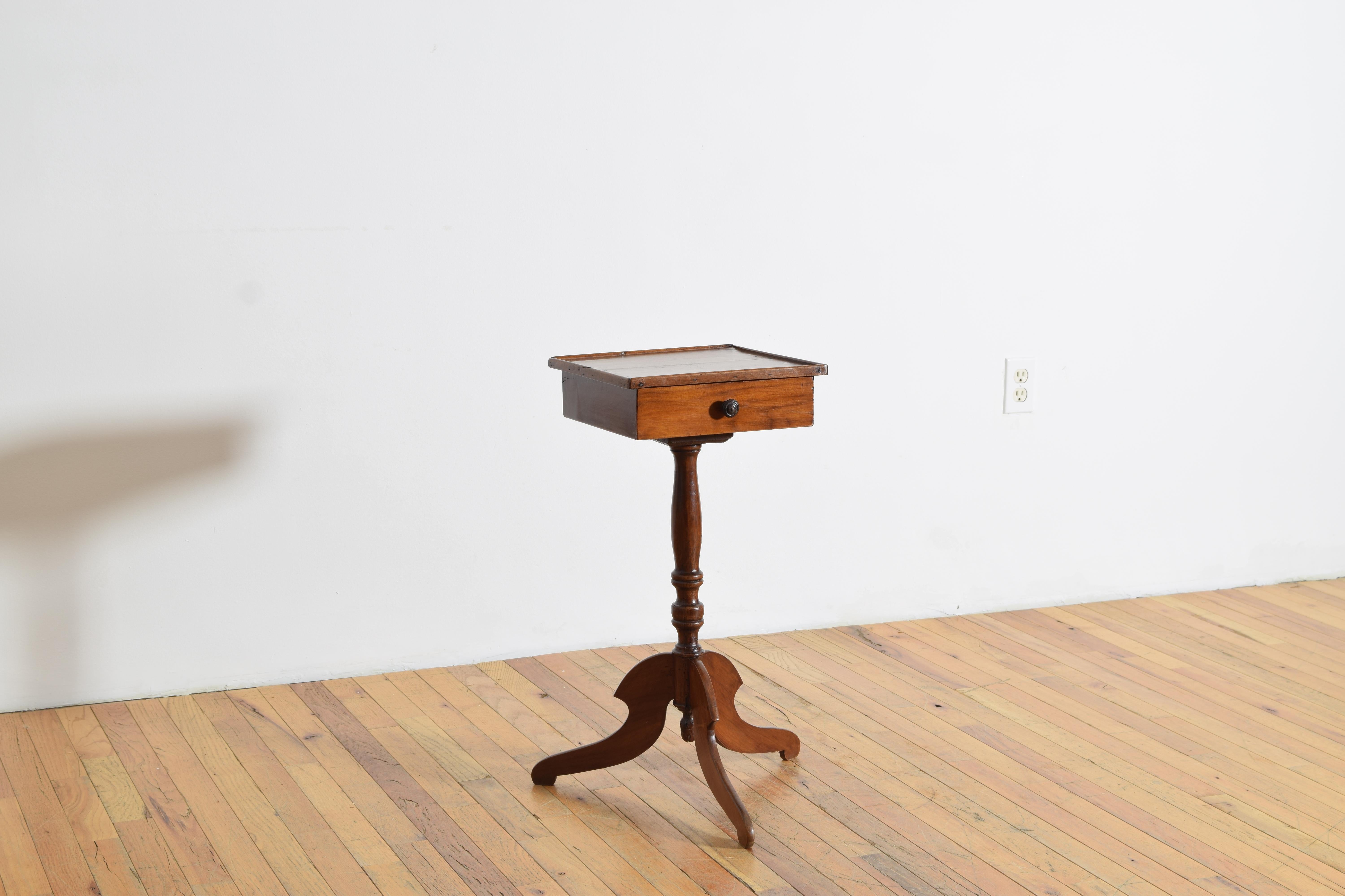 Having a square top with a slightly raised gallery atop a conforming case housing a single drawer, the top raised on a turned standard atop a tripartite base issuing shaped legs, diameter of base 10”