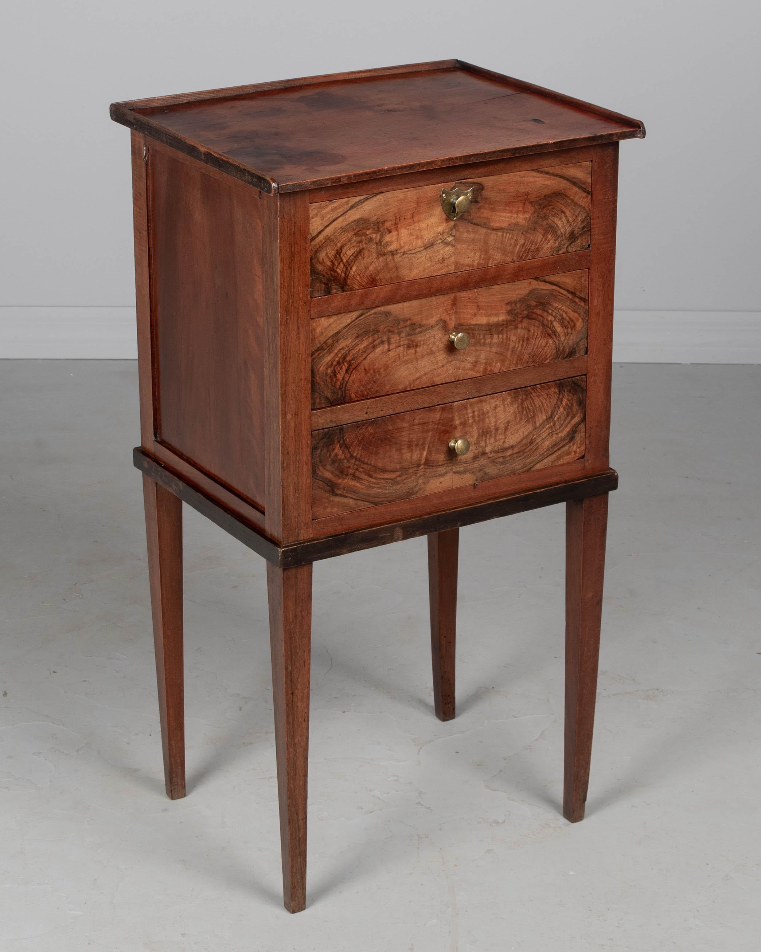 Mesa auxiliar o de noche francesa de nogal Louis Philippe de principios del siglo XX. Bonitas vetas de madera en la cara de los cajones. Acabado por los cuatro costados. Tres cajones con pequeños pomos y escudo de latón. Robustas y esbeltas patas