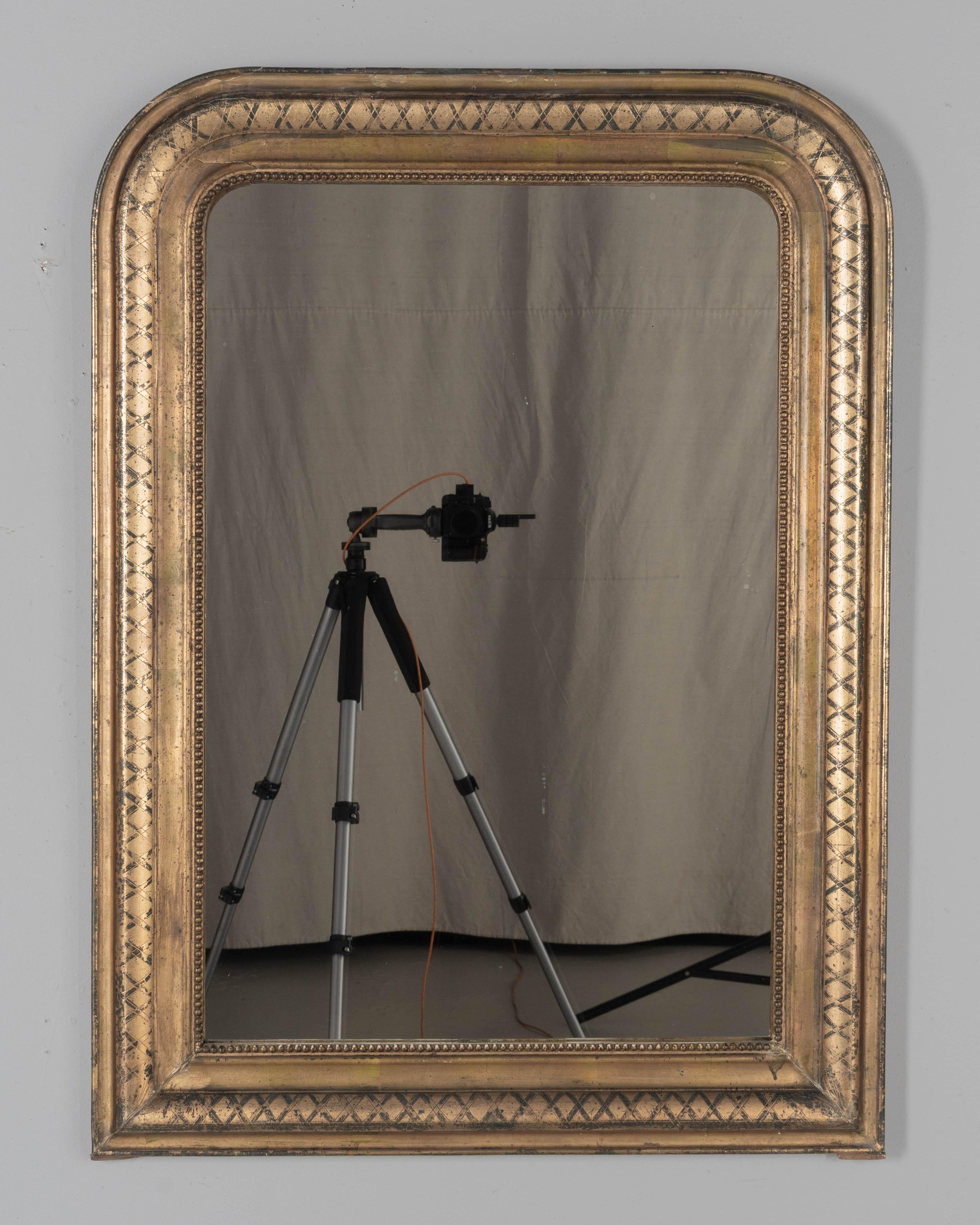 Espejo dorado de estilo Louis Philippe francés de principios del siglo XX, con esquinas superiores curvadas, motivo de x incisa y borde interior de cuentas. Bonita pátina dorada con el negro del roce al descubierto. El espejo original tiene ligeros